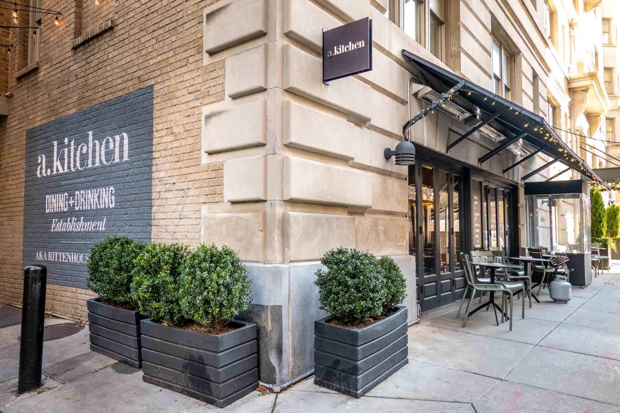 Corner of a building with outdoor seating on one side and a sign on the other side: "A.kitchen, dining and drinking establishment, AKA Rittenhouse"