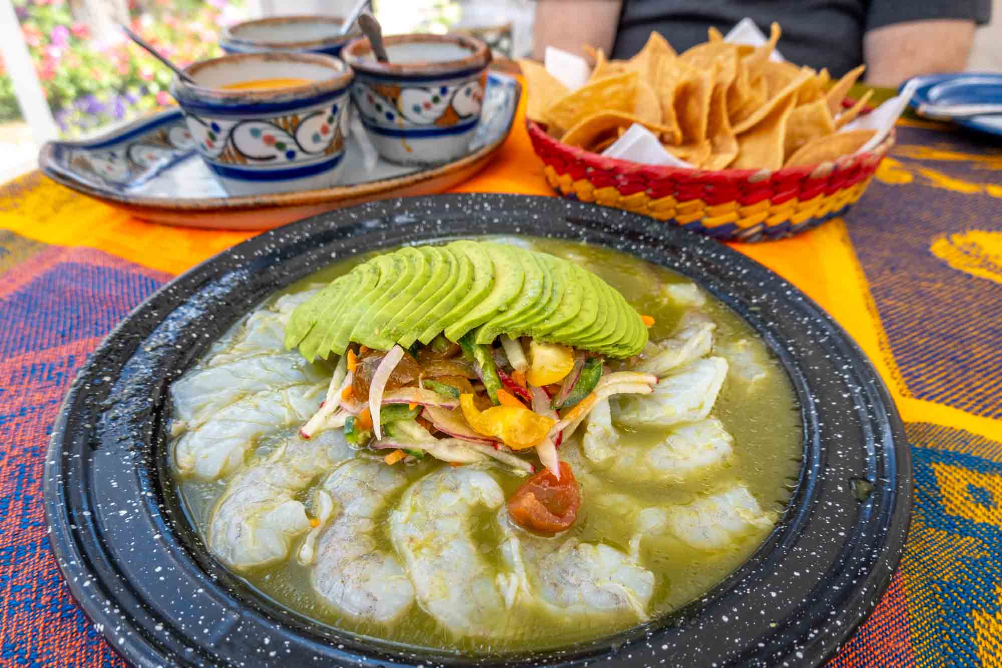 Plate of shrimp aquachile with avocado and basket of tortilla chips in the background.