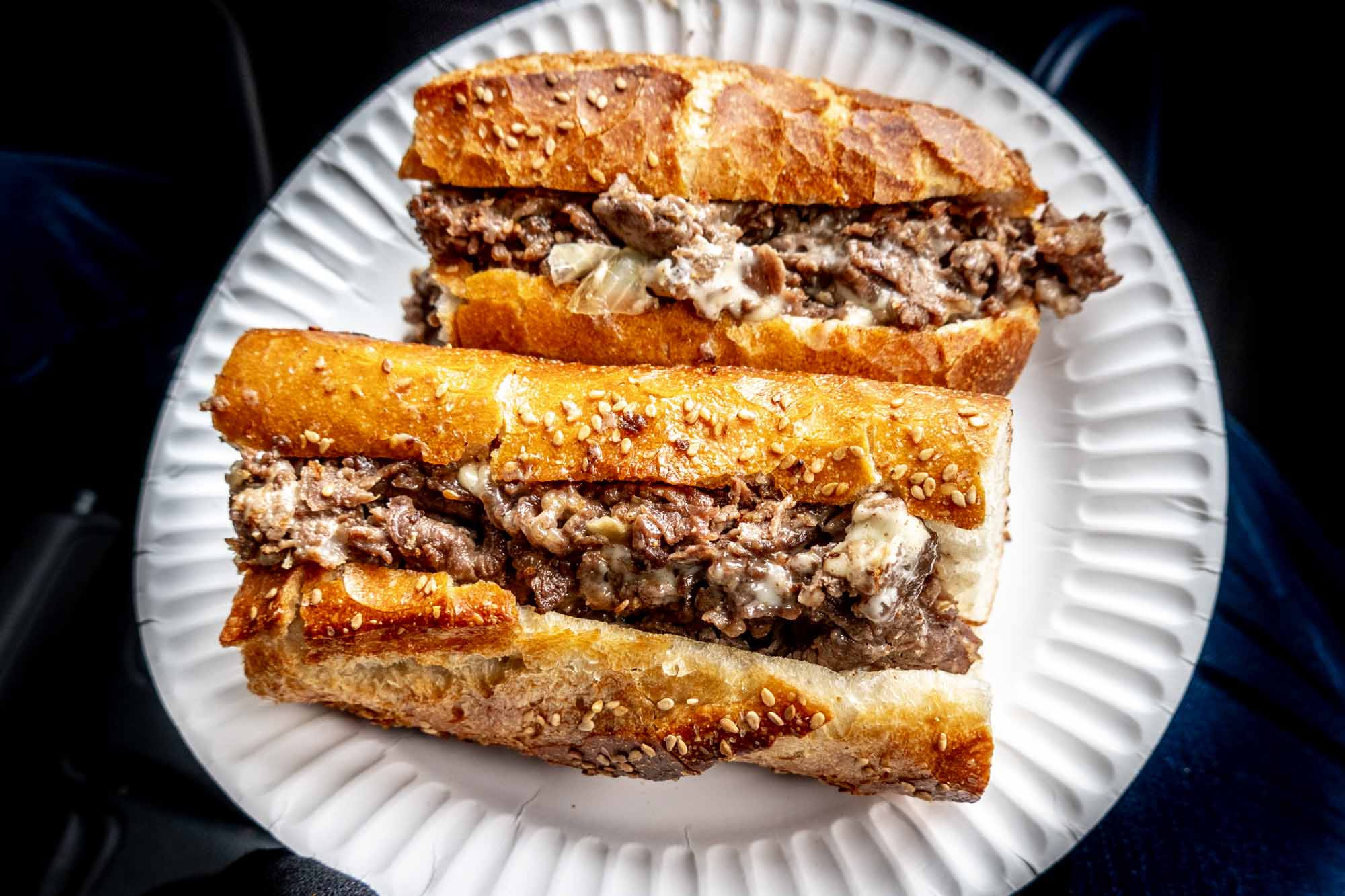 Cheesesteak cut in half on white paper plate.