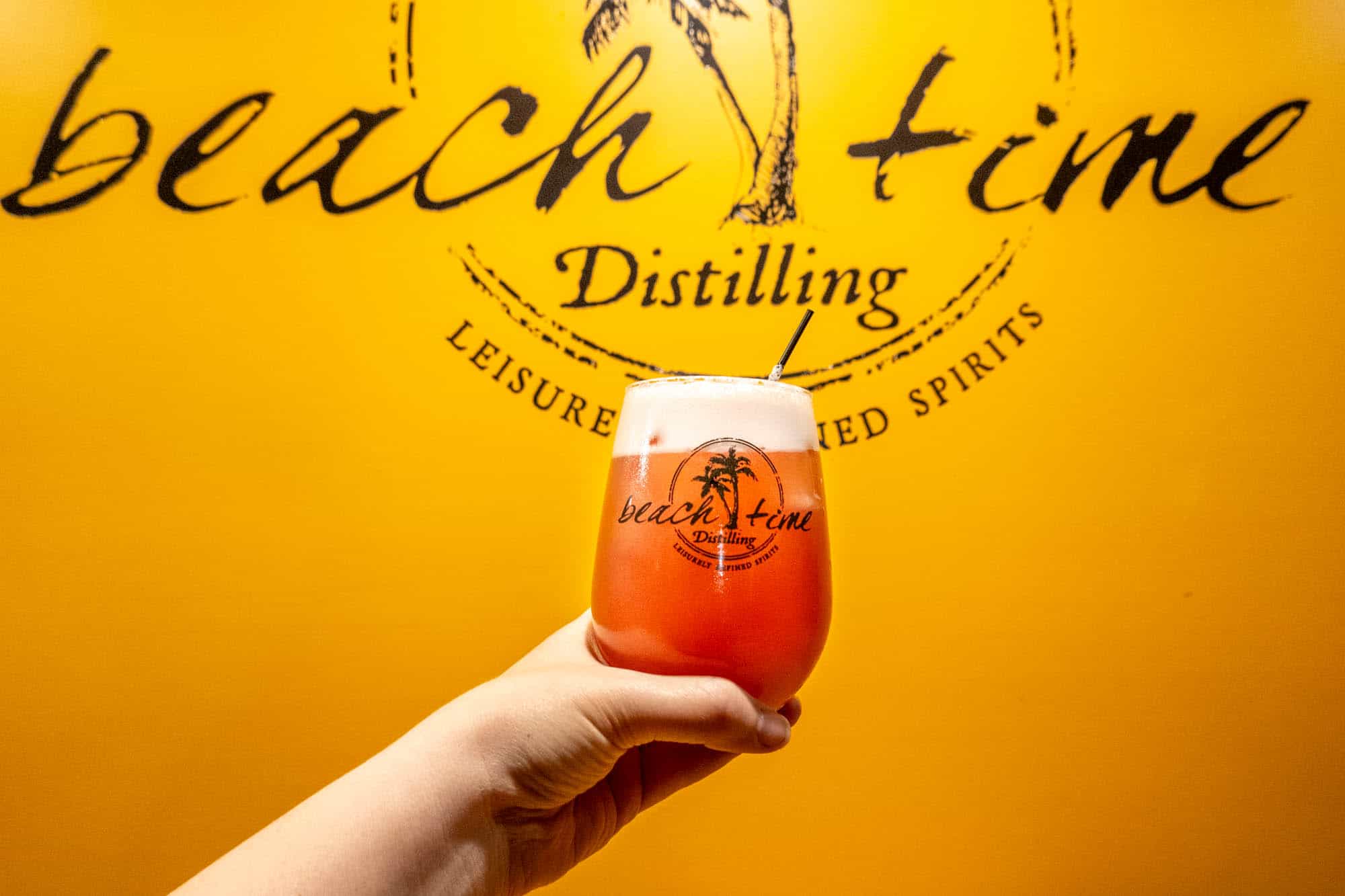Hand holding a cocktail in front of a sign for "Beach Time Distilling"