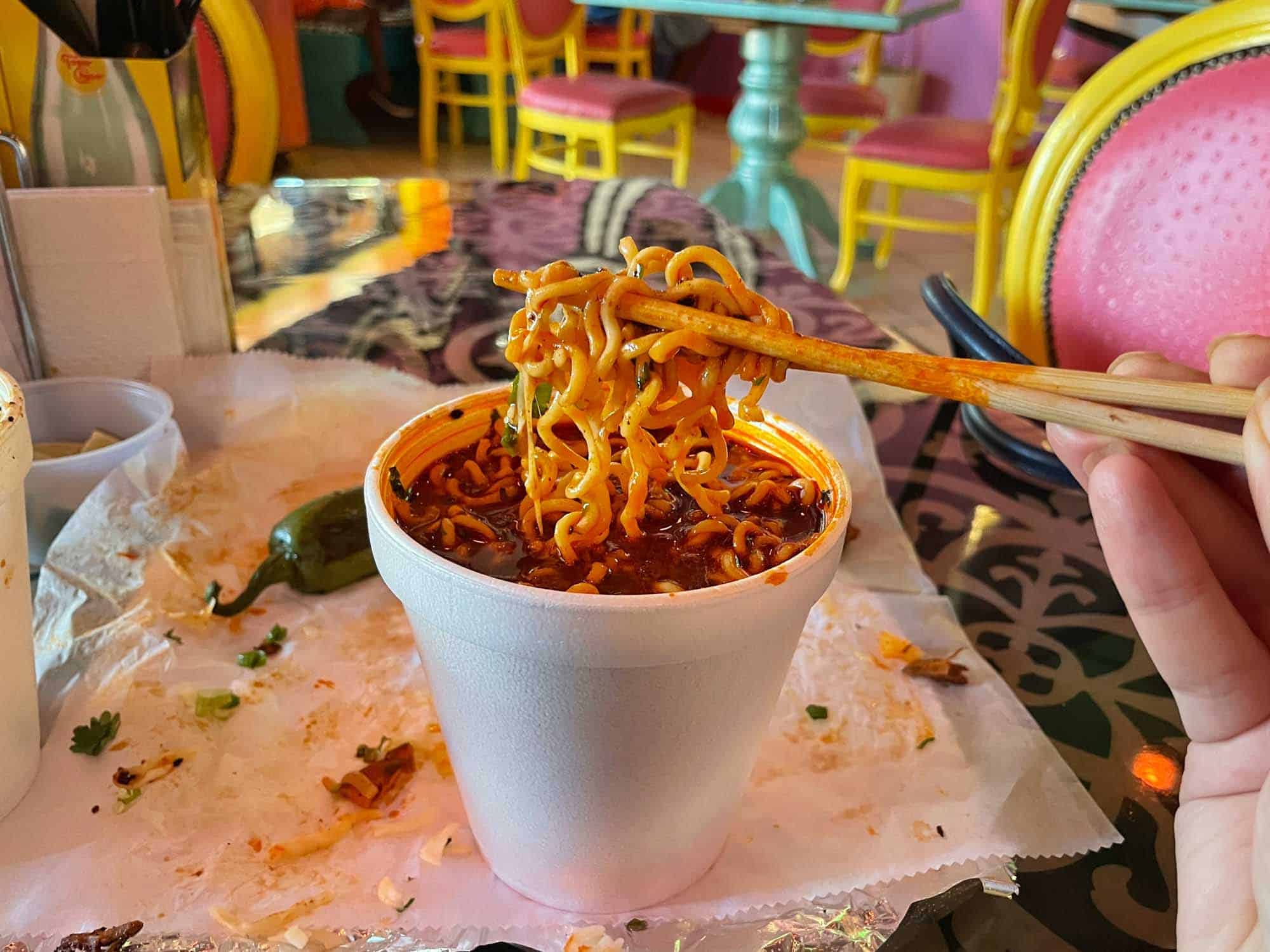 Chopsticks holding noodles above a cup of birria ramen.