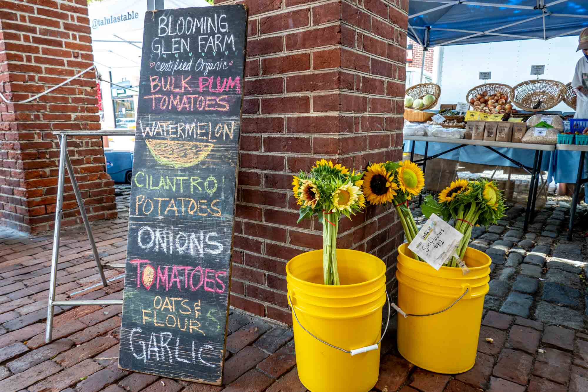 Yellow buckets of sunflowers next to a board offering fresh vegetables at an outdoor market