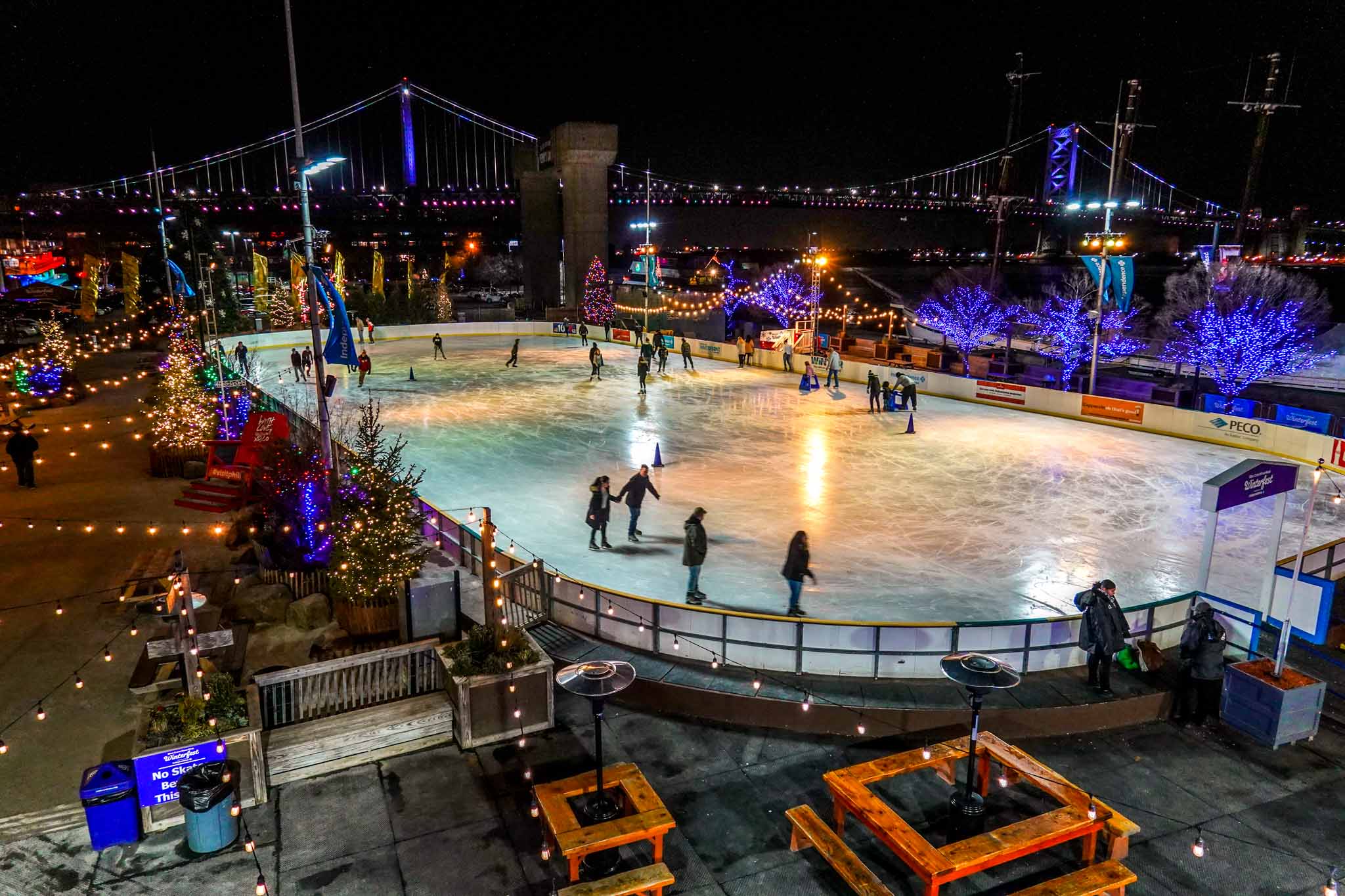 Overhead view of skaters on an ice-skating rink surrounded by Christmas lights at night.