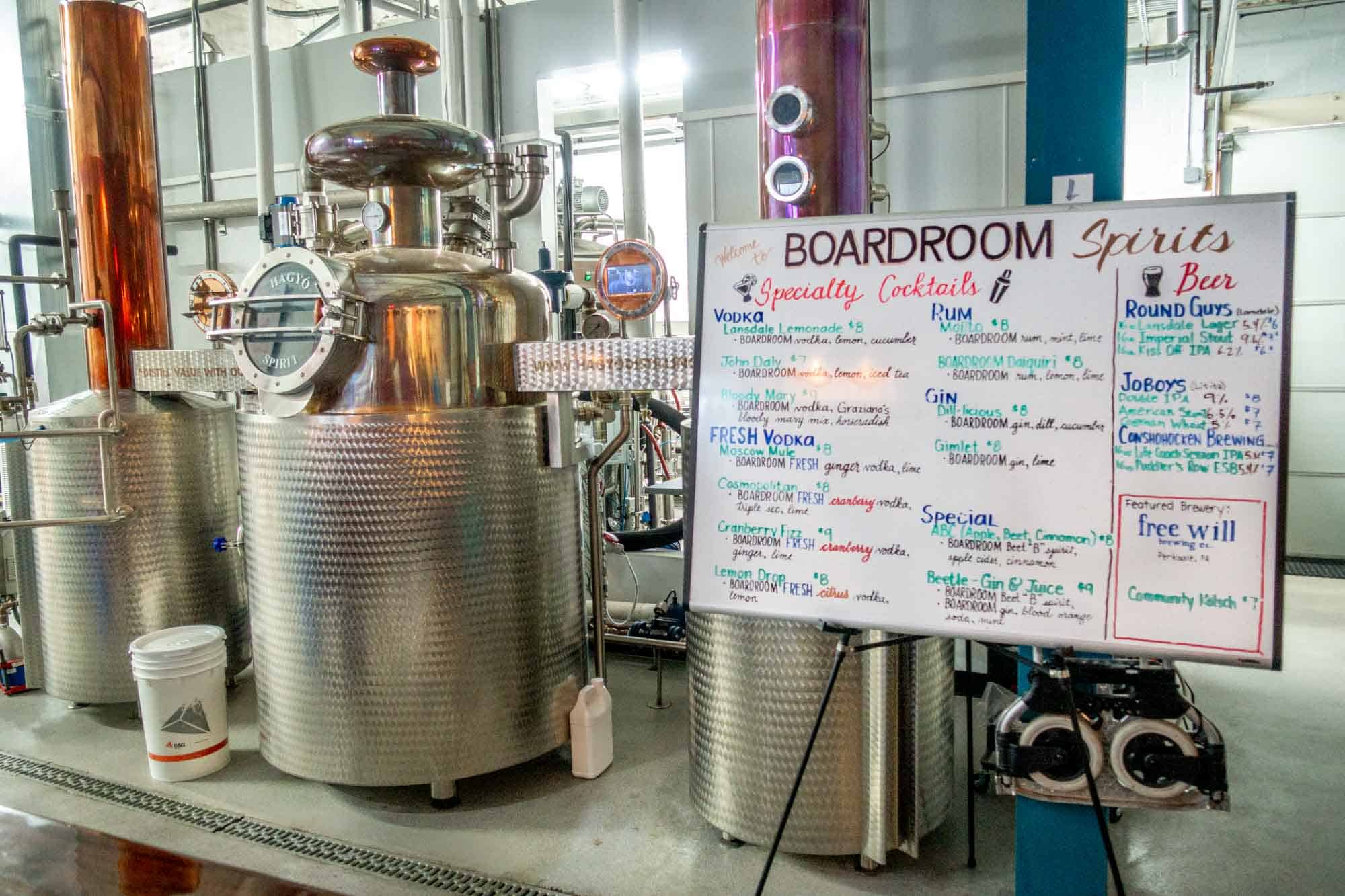 Copper stills in a distillery beside a cocktail menu written on a whiteboard