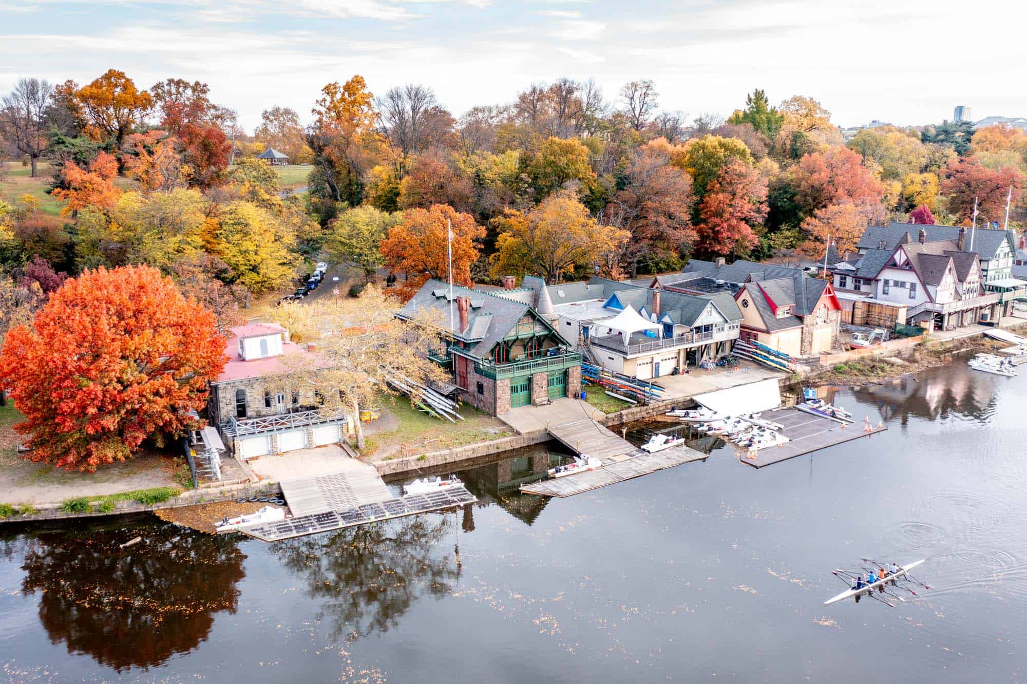 Crew rowing in the Schuylkill River in front of a row of boathouses and trees with colorful leaves. 