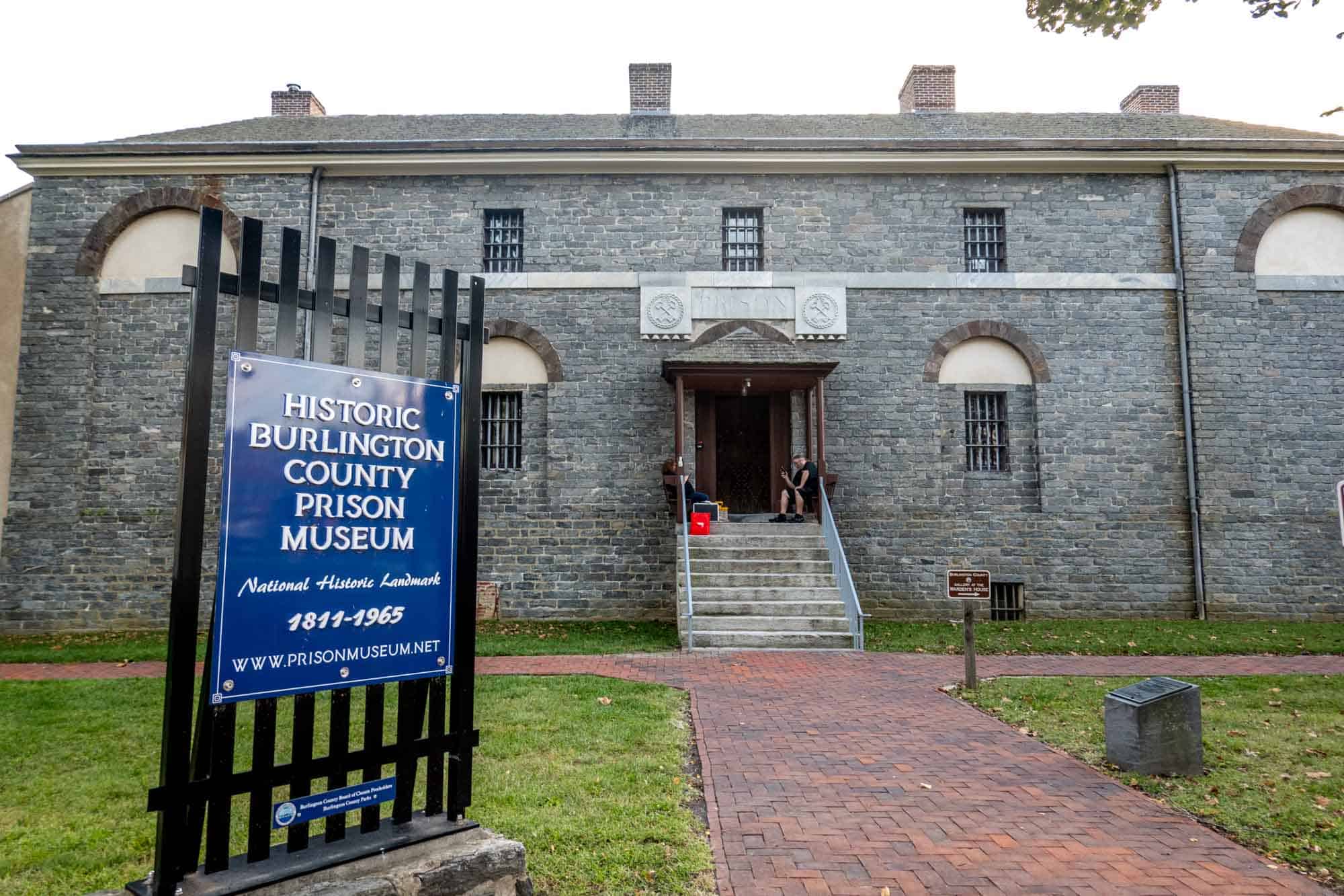 Gray brick building with barred windows behind a sign: "Historic Burlington County Prison Museum"