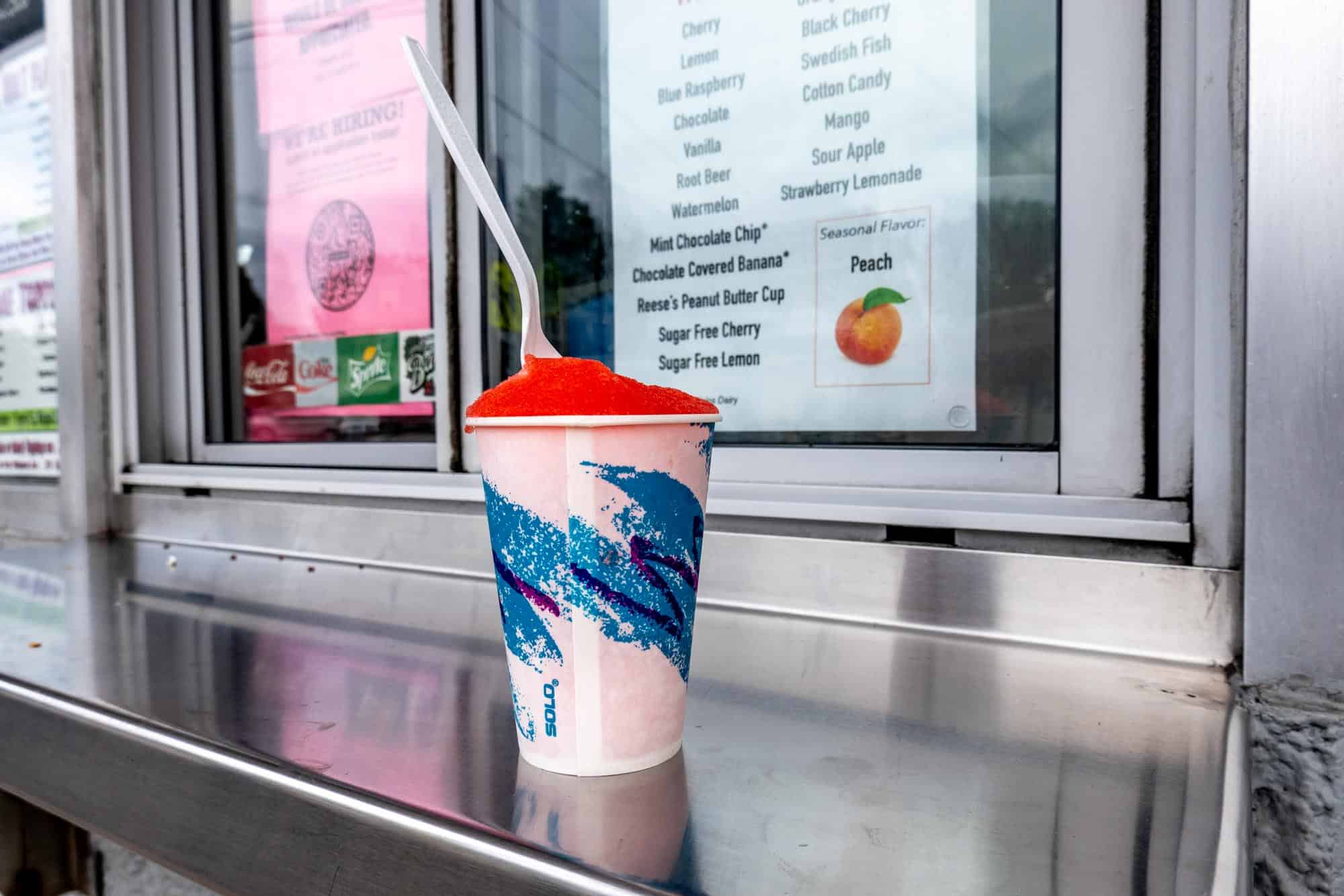 Cup of red water ice with a spoon in it on a steel window ledge outside a service window.