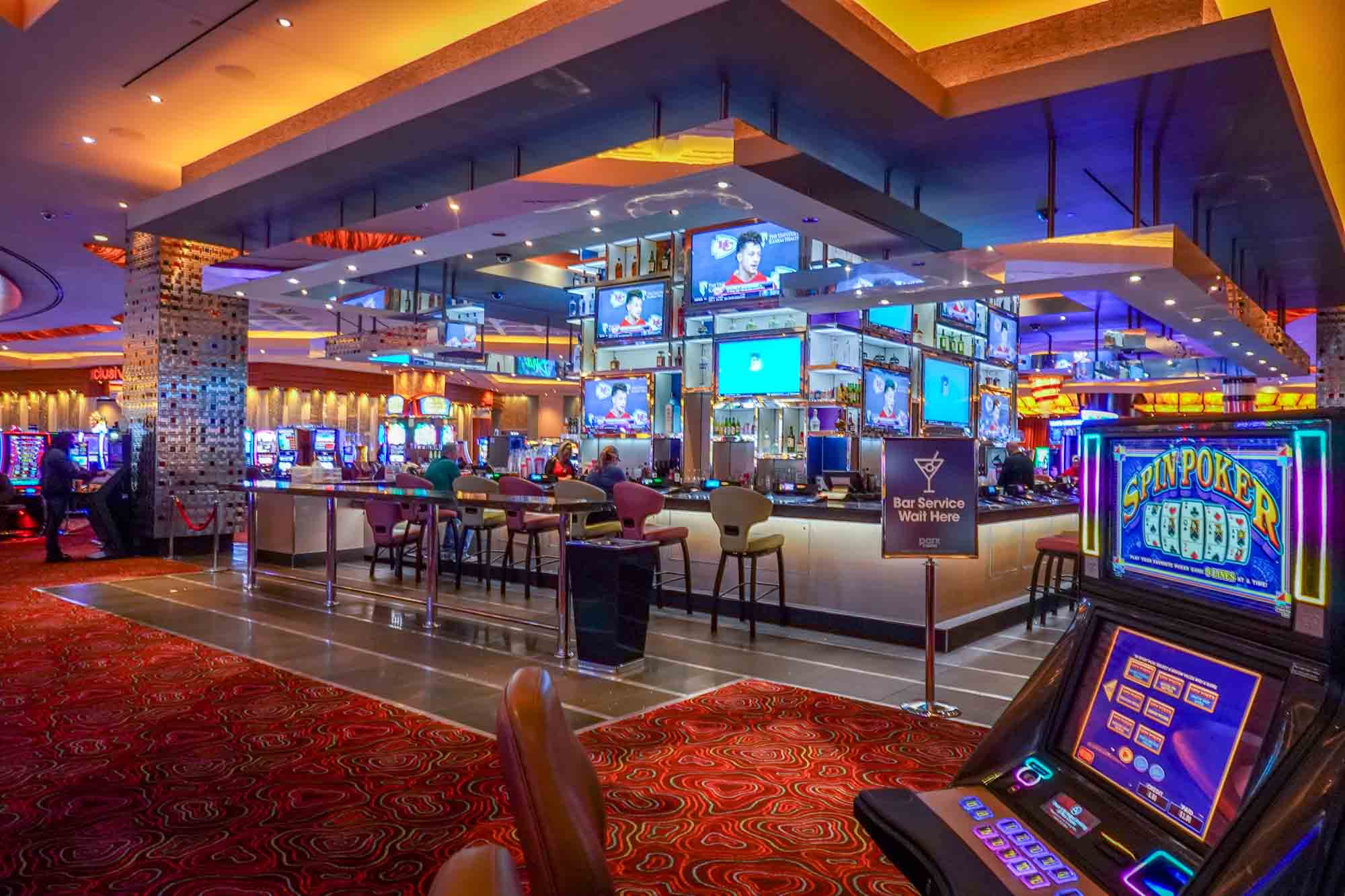 Bar with numerous TVs surrounded by electronic gambling games on a casino floor. 