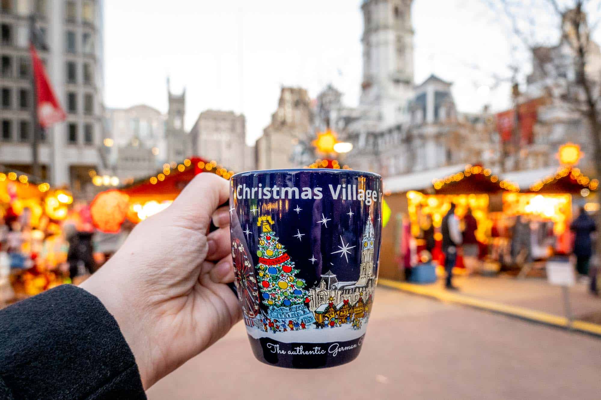 Hand holding a blue mug labeled "Christmas village" in front of Christmas lights.