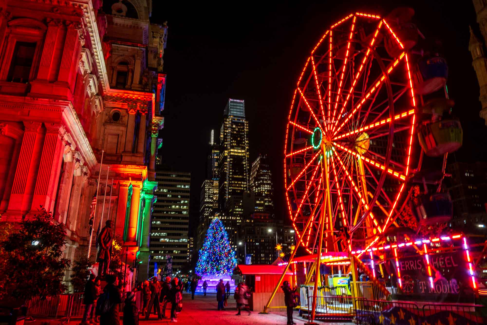 Ferris wheel lit up with red lights at night with a lit up Christmas tree and city buildings in the background