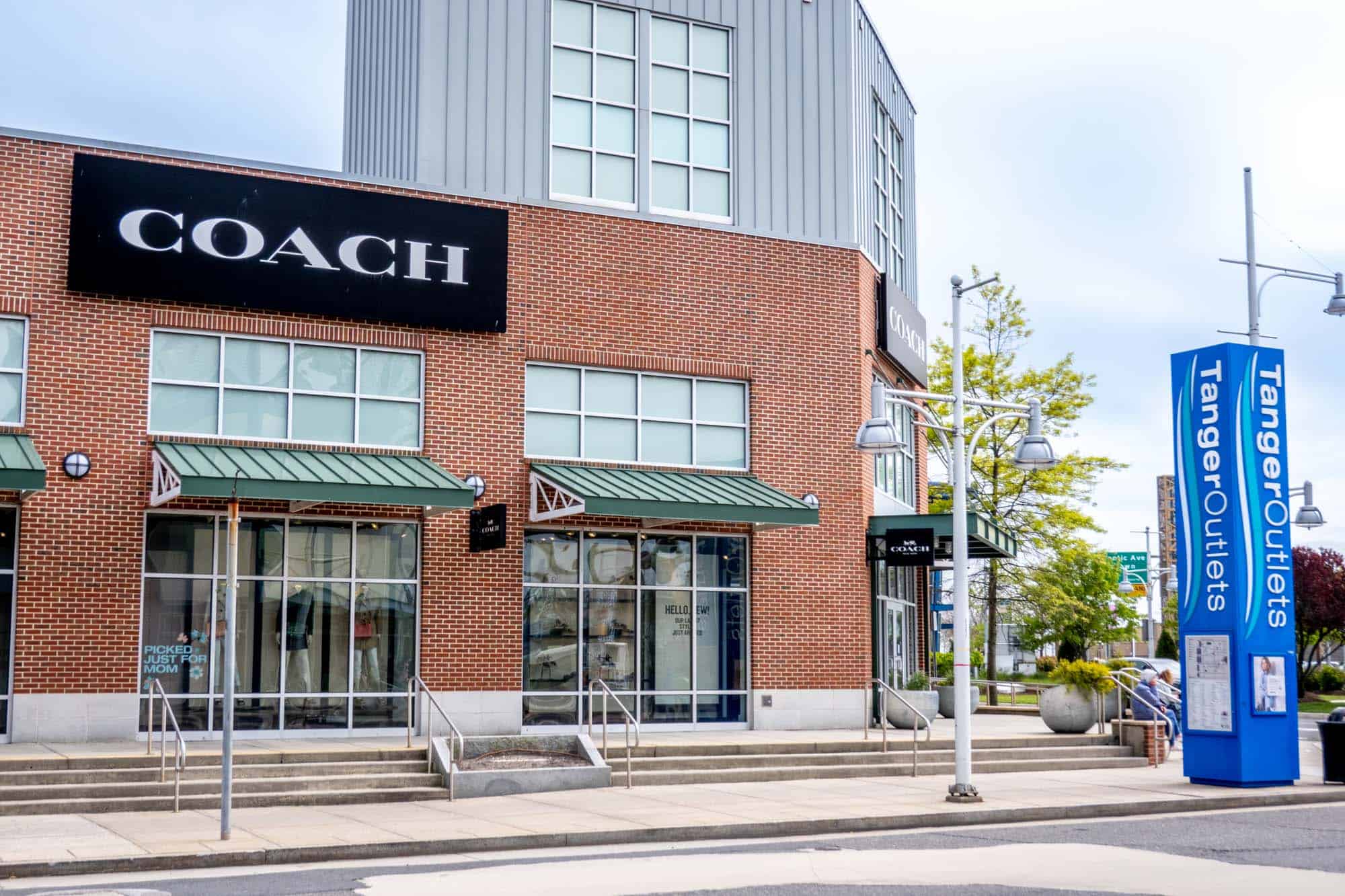 Brick building with green awnings and a sign for "Coach" next to a blue pillar with a sign for "Tanger Outlets"