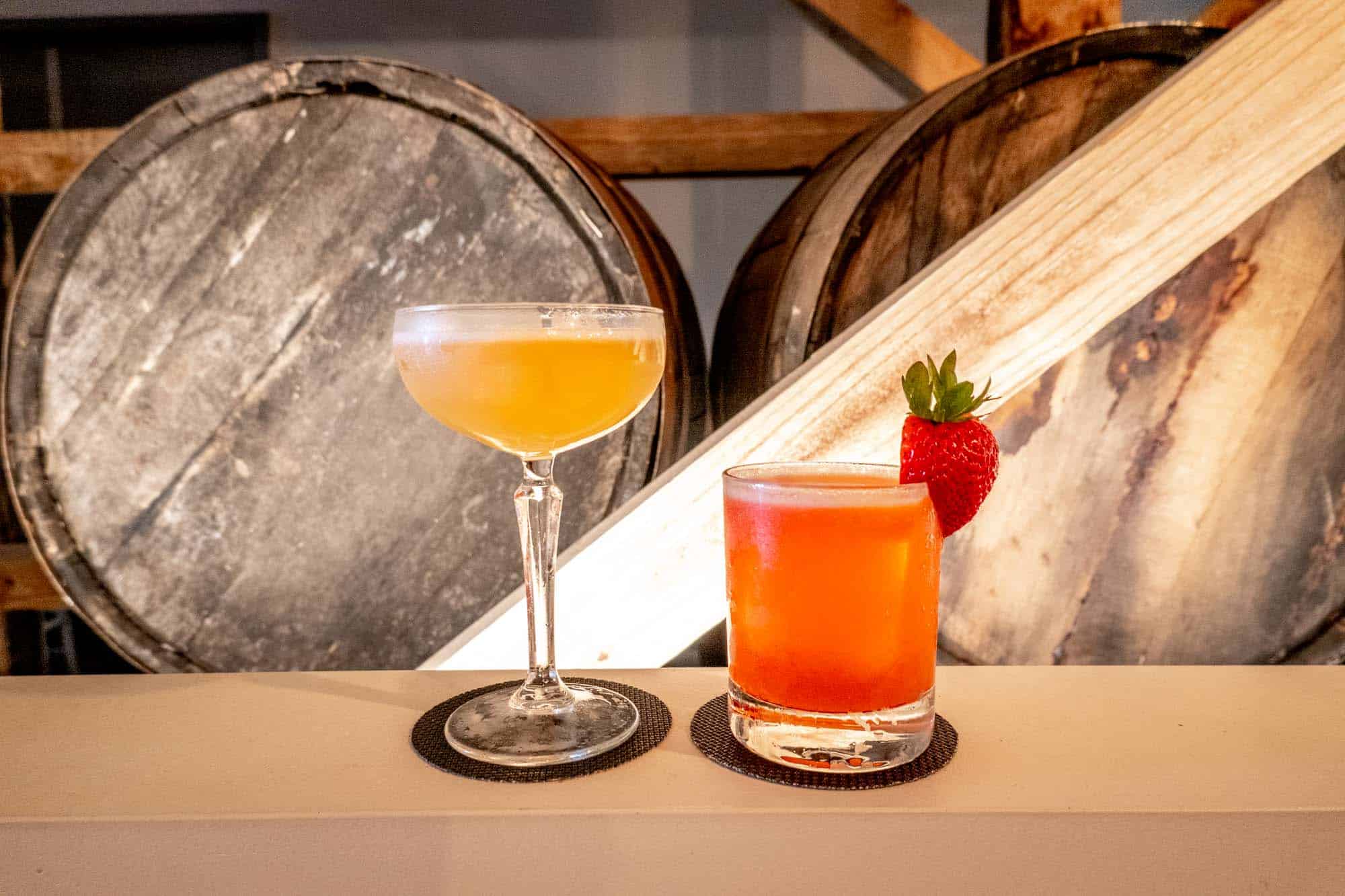 Two cocktails on a shelf in front of two wooden whiskey barrels
