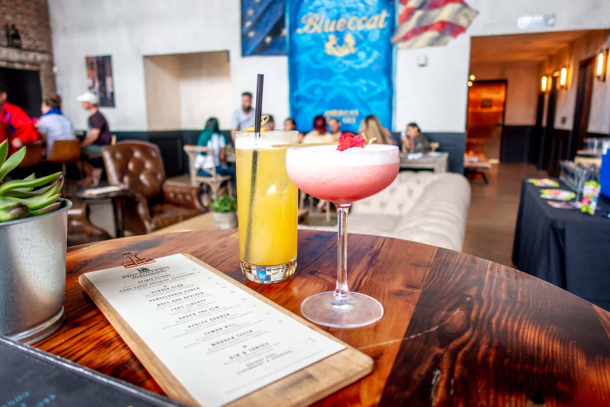 Two cocktails and a menu on a table in front of a mural showing a bottle of Bluecoat gin