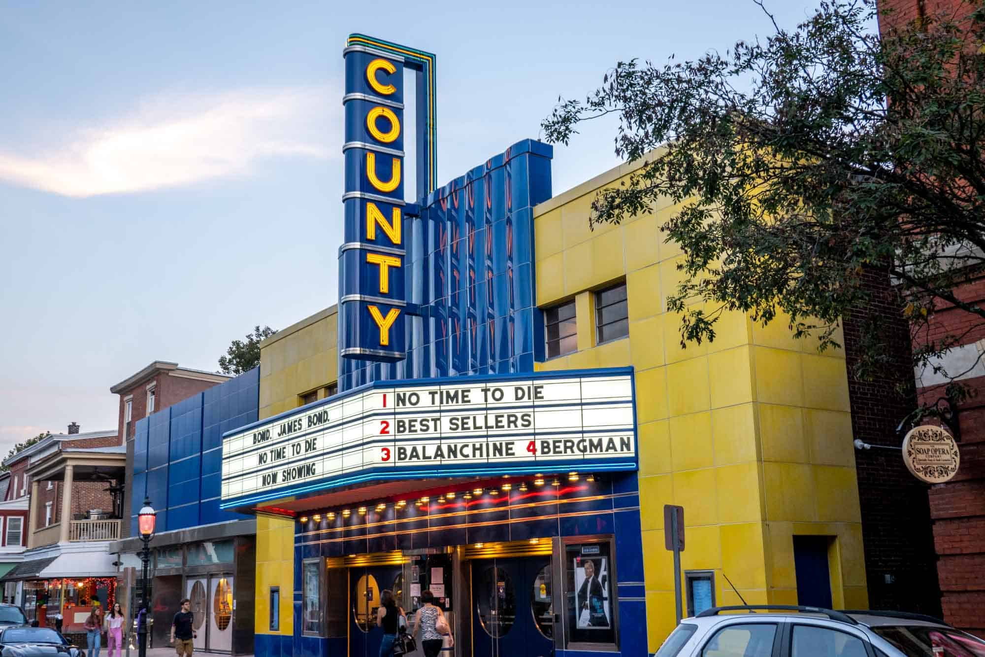 Exterior of a movie theater with a marquis advertising movies and a large blue sign: "County"