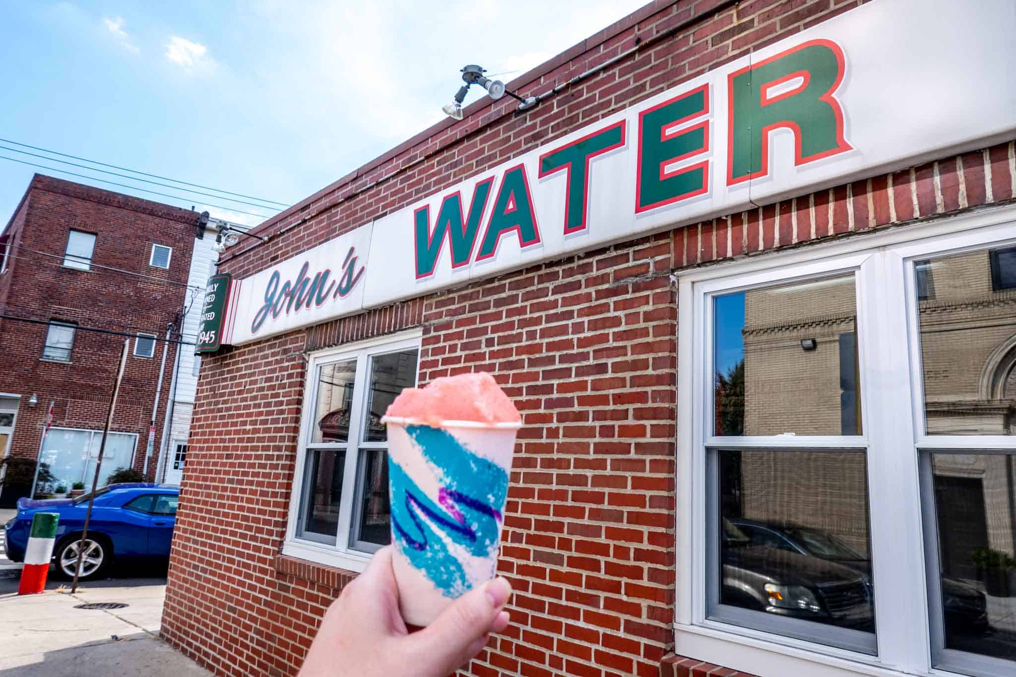 Cup filled with cherry water ice in front of a sign for John's.