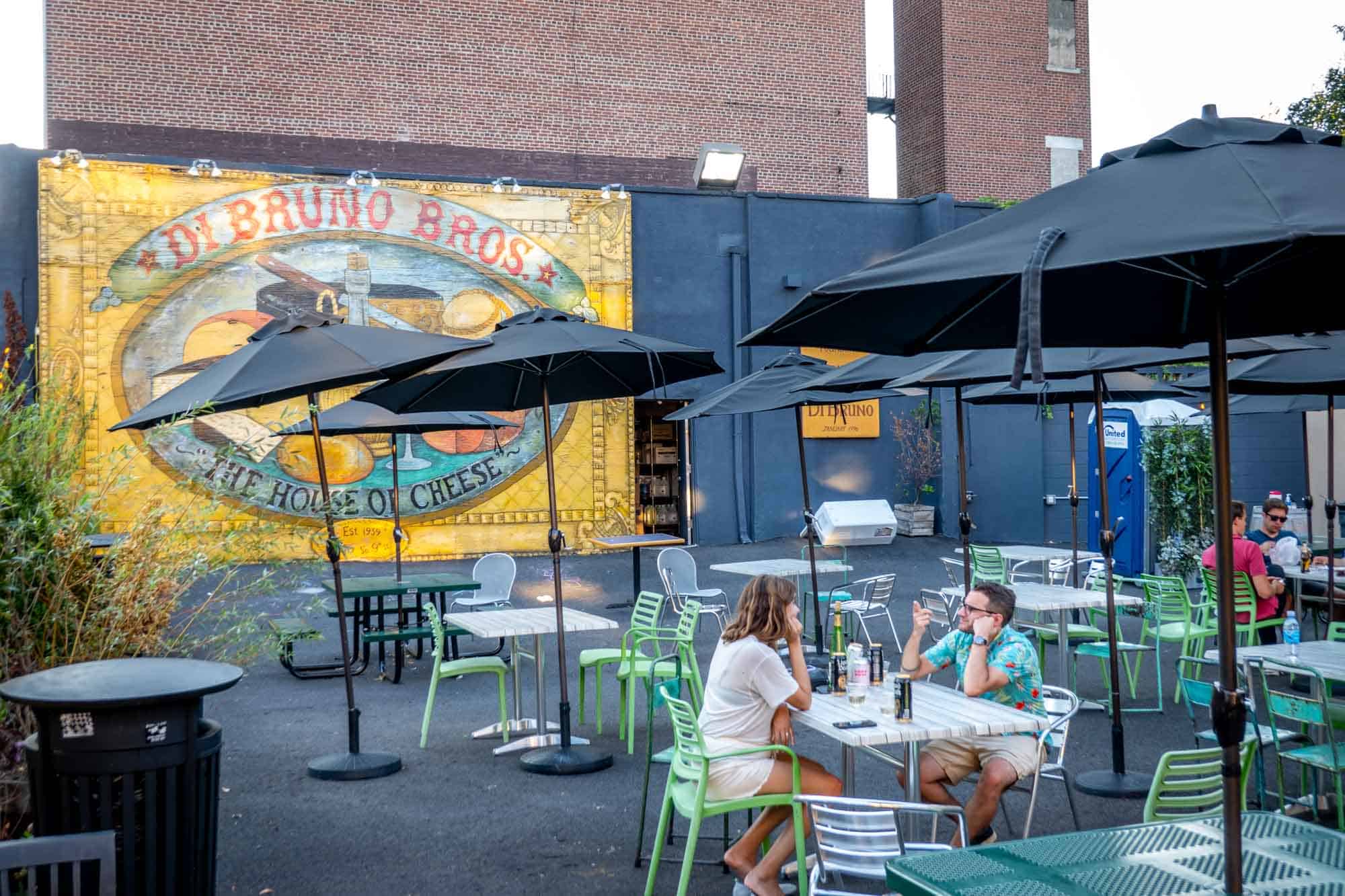 People drinking at picnic tables under umbrellas in front of DiBruno Bros mural.