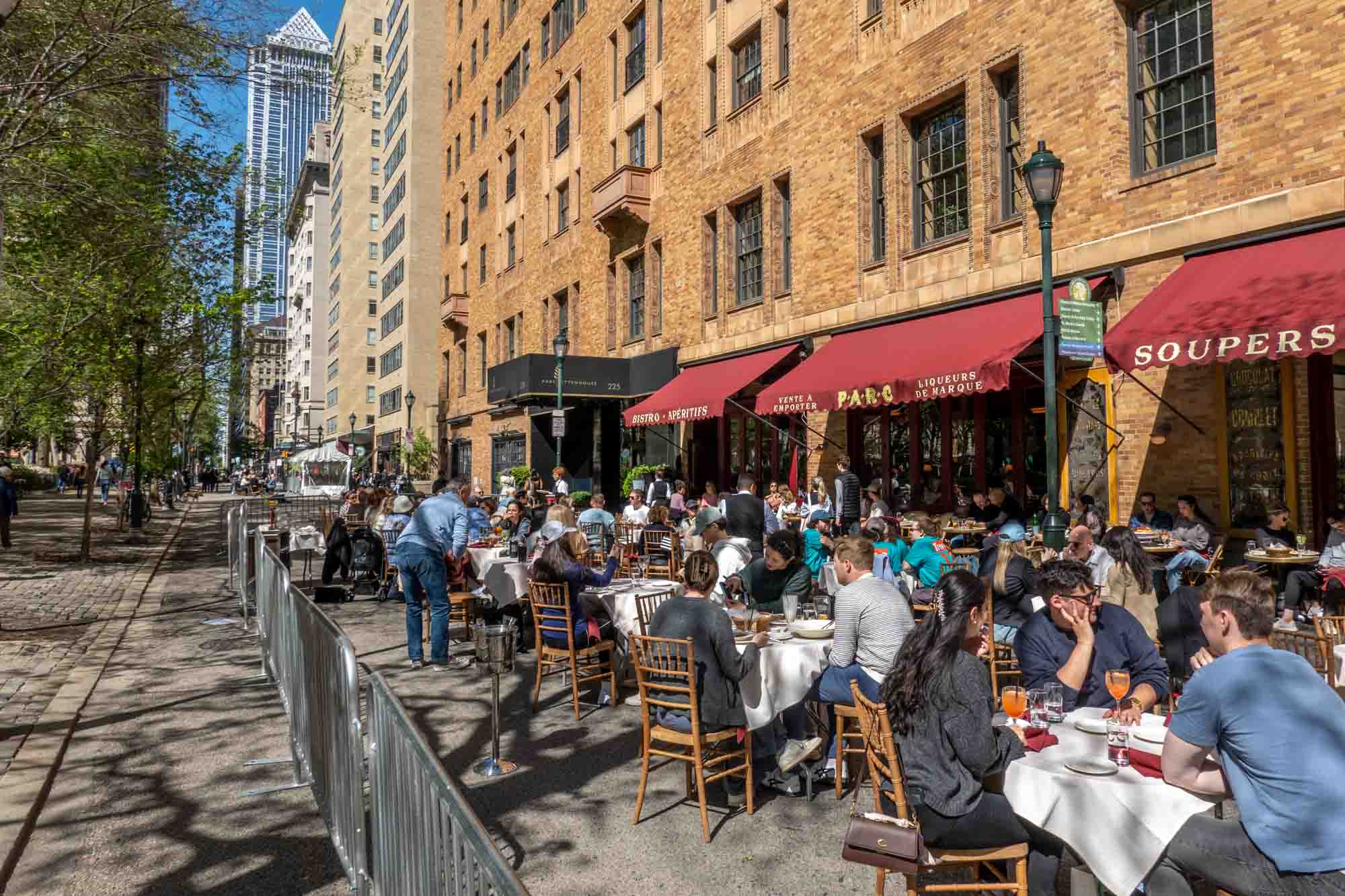 People sitting at outdoor restaurant tables that spill into the street on a sunny day.