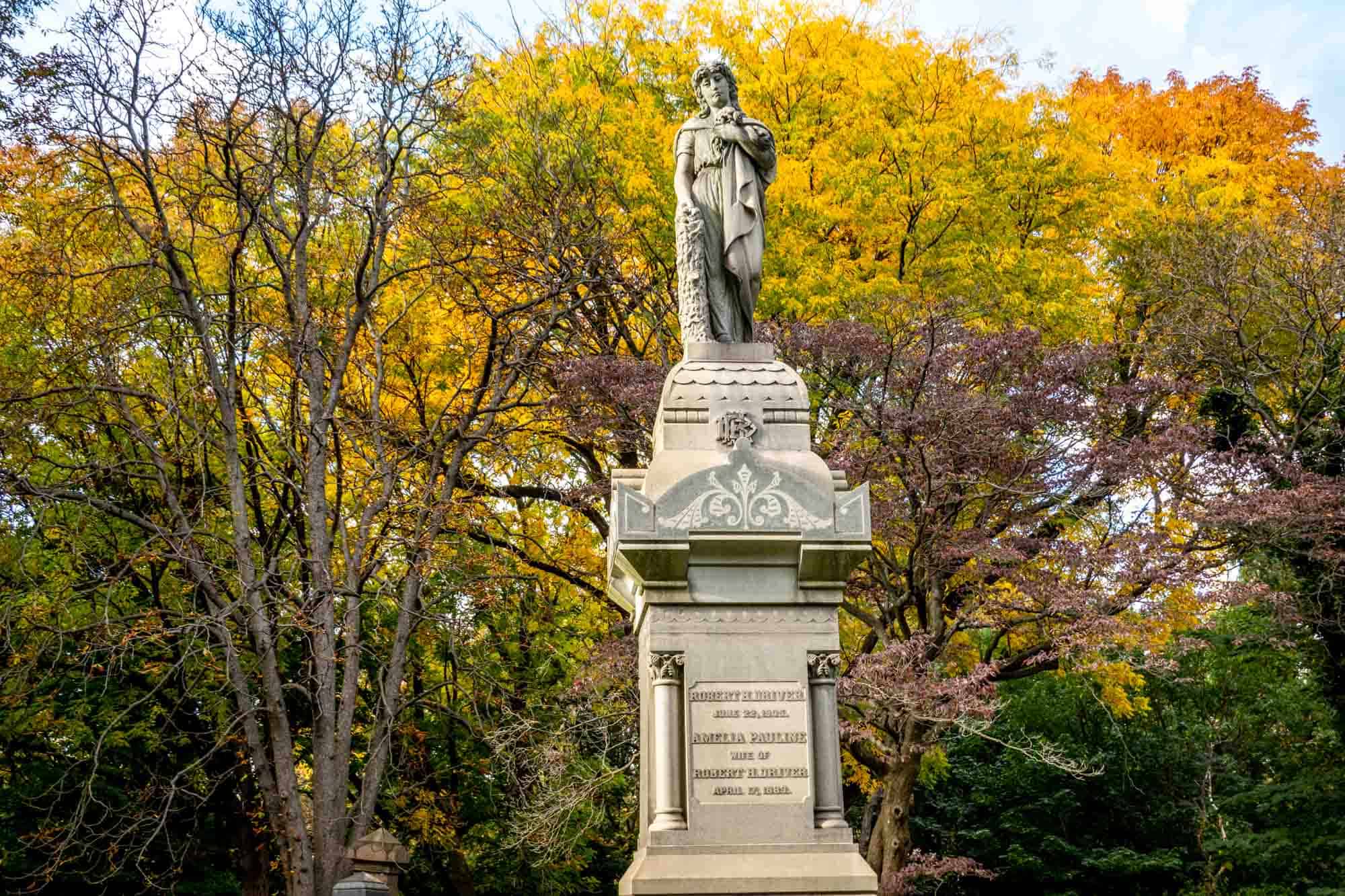 Large grave marker with the statue of a person standing on an elaborate platform in front of trees with bright fall leaves
