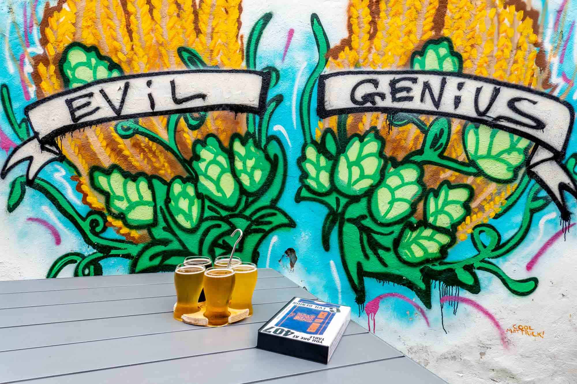 Flight of beer glasses on a table in front of a mural of wheat labeled "Evil Genius."