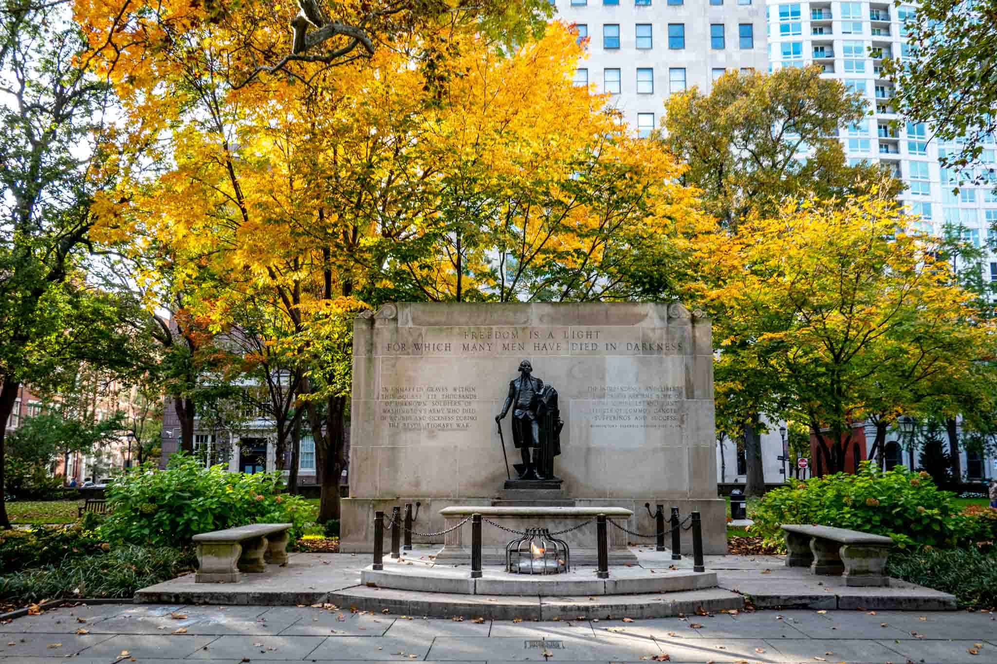 Monument with a statue of a man and an eternal flame in front of a tree with yellow leaves.