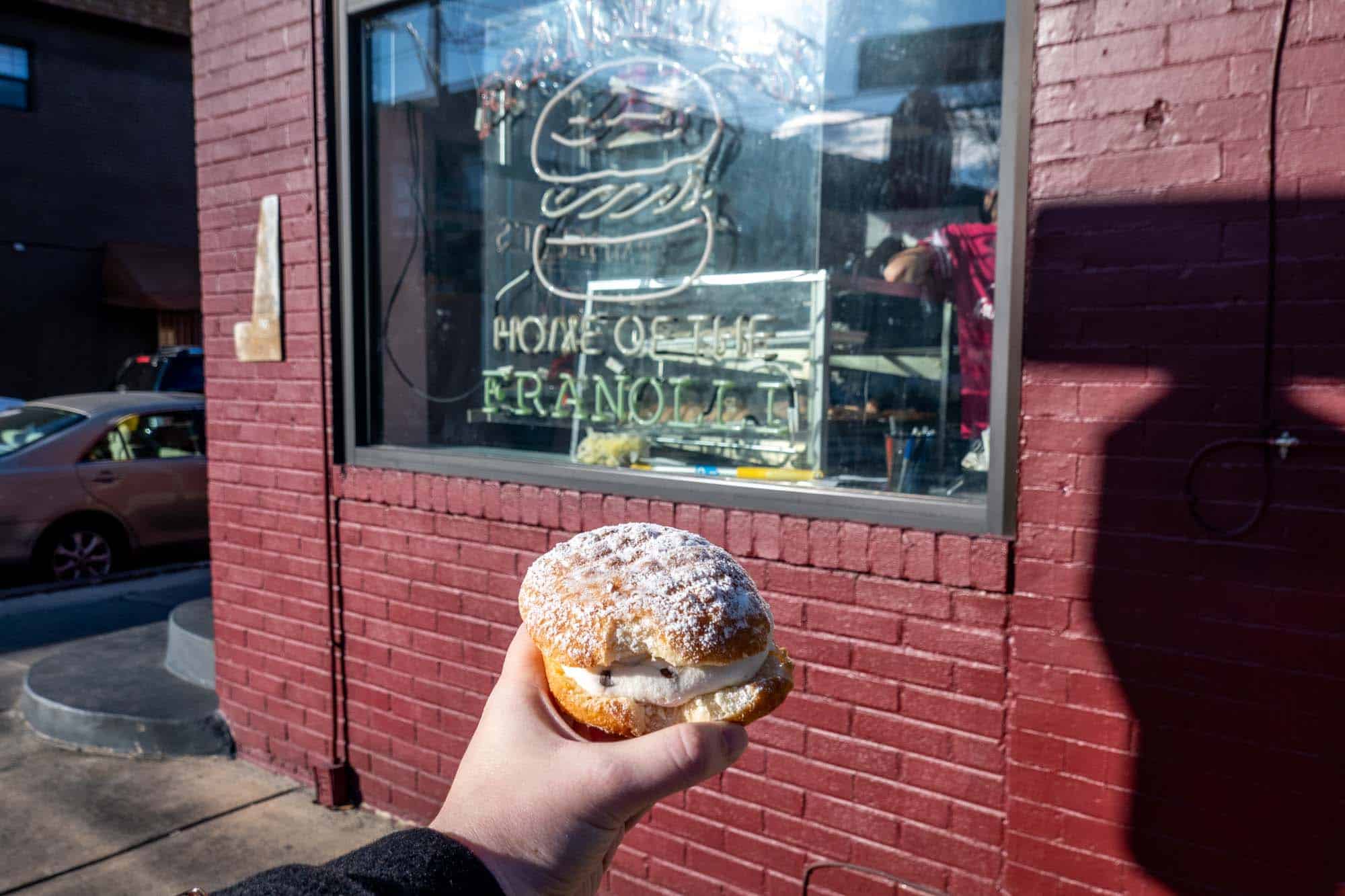 Hand holding a pastry in front of a sign for "Frangelli's: Home of the Franolli."