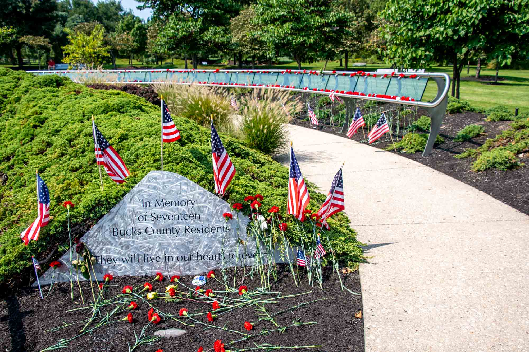 American flags and flowers around a stone slab engraved with: "In Memory of Seventeen Bucks County Residents--They will live in our hearts forever"