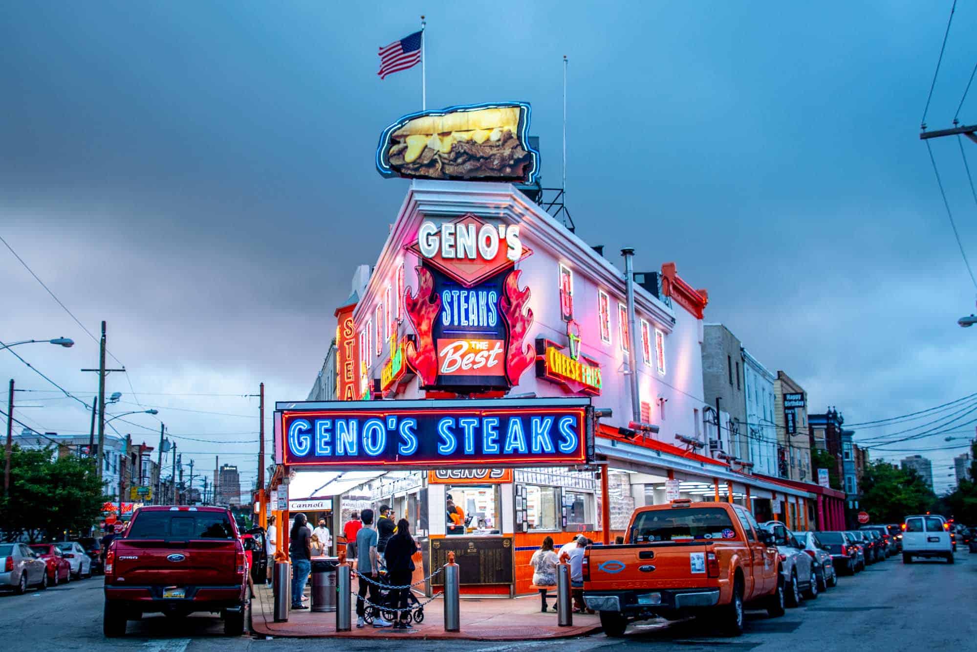 Exterior of a corner restaurant with customers outside and a sign for "Geno's Steaks."