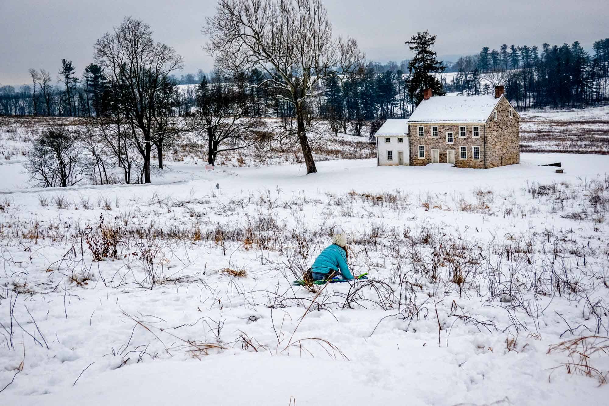 Girl in turquoise jacket sledding toward a stone building