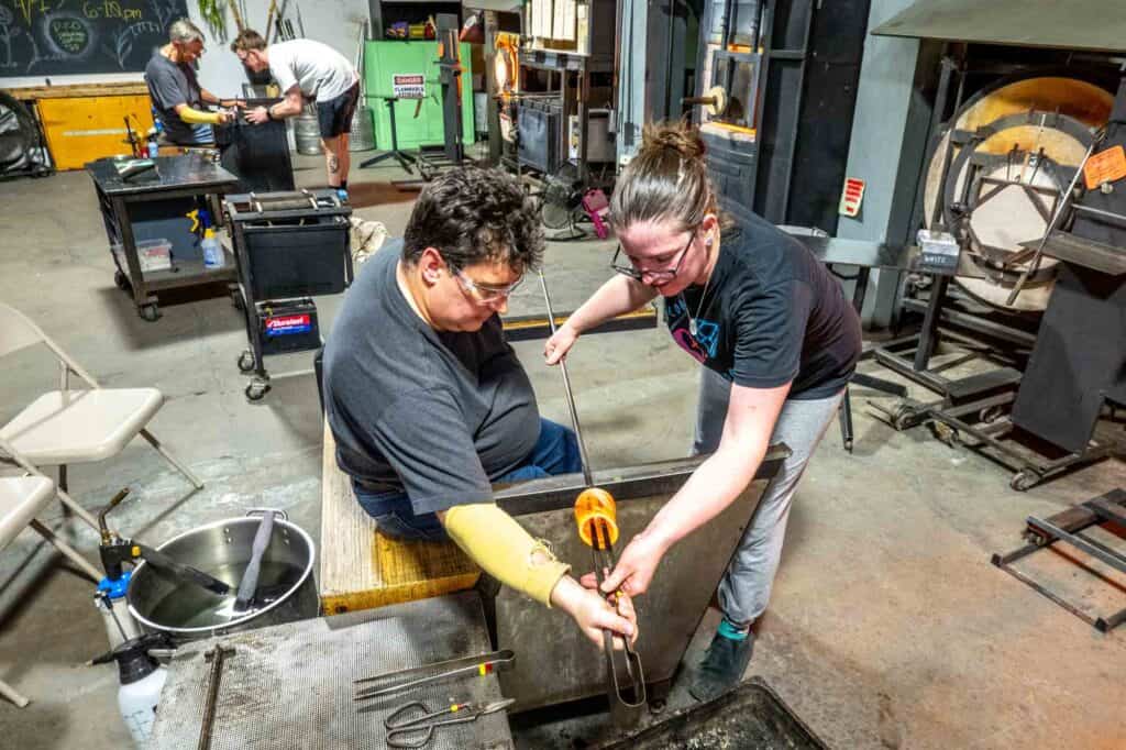 Two people creating a drinking glass in a glass blowing workshop.