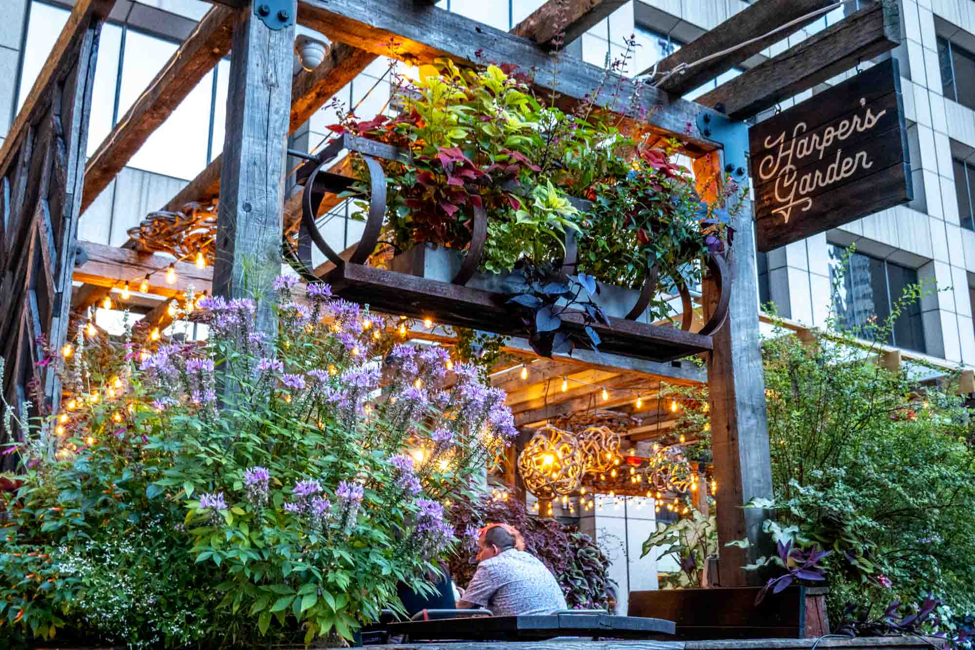 Restaurant patio filled with plants, flowers, and warm light fixtures and a sign for "Harper's Garden"
