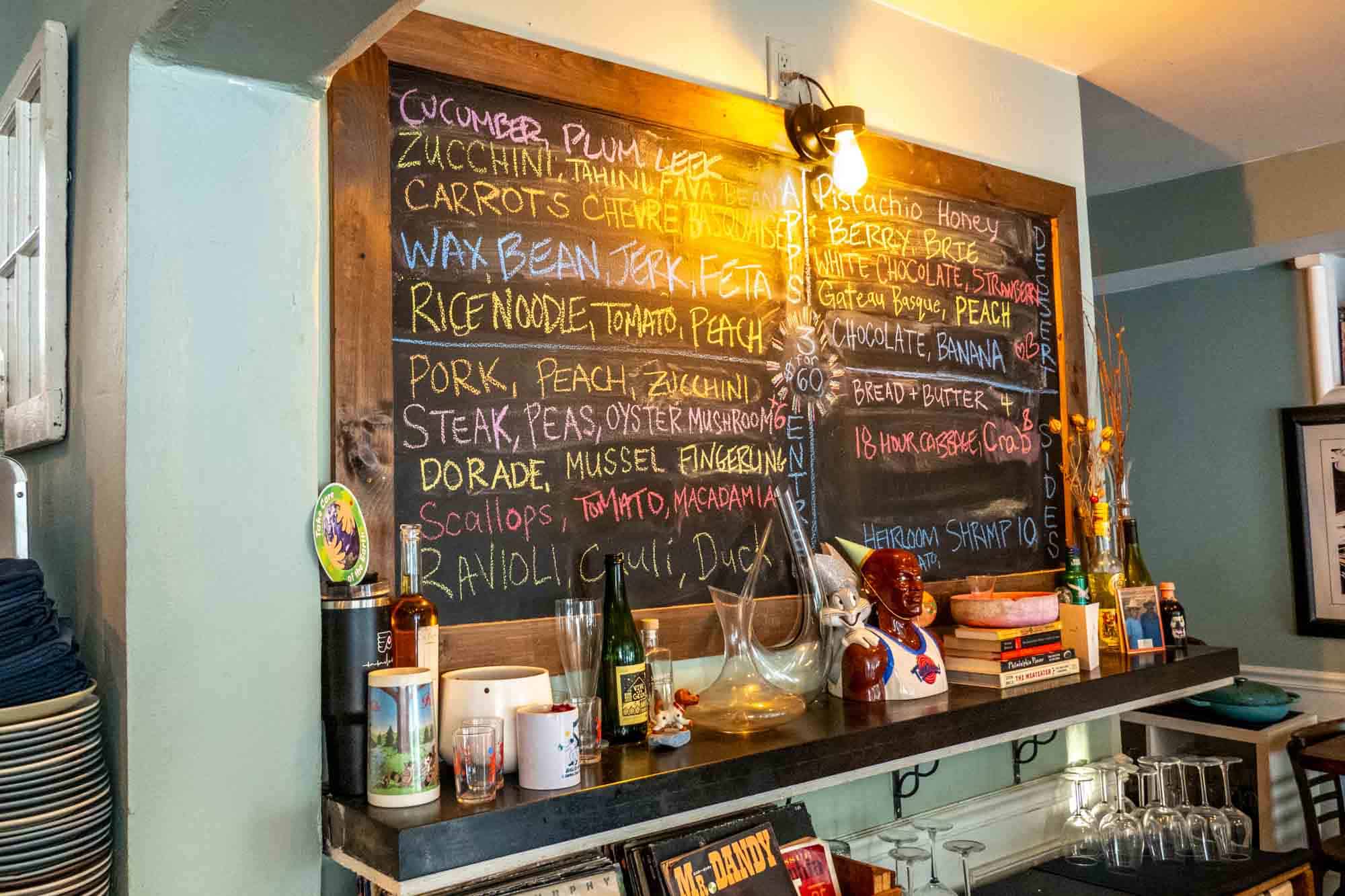 Blackboard menu on the wall of a restaurant above a shelf filled with dishes and knickknacks. 