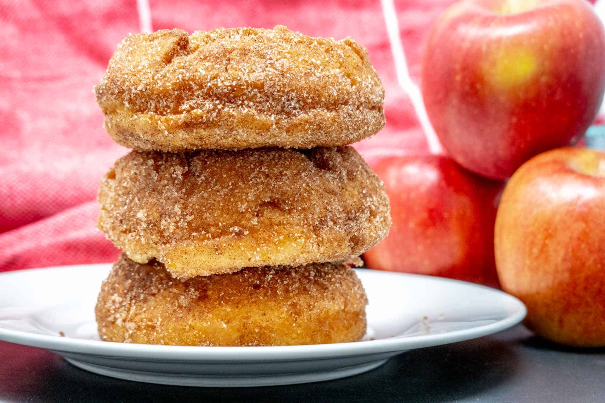 Three cinnamon-sugar donuts stacked on a plate beside a stack of apples