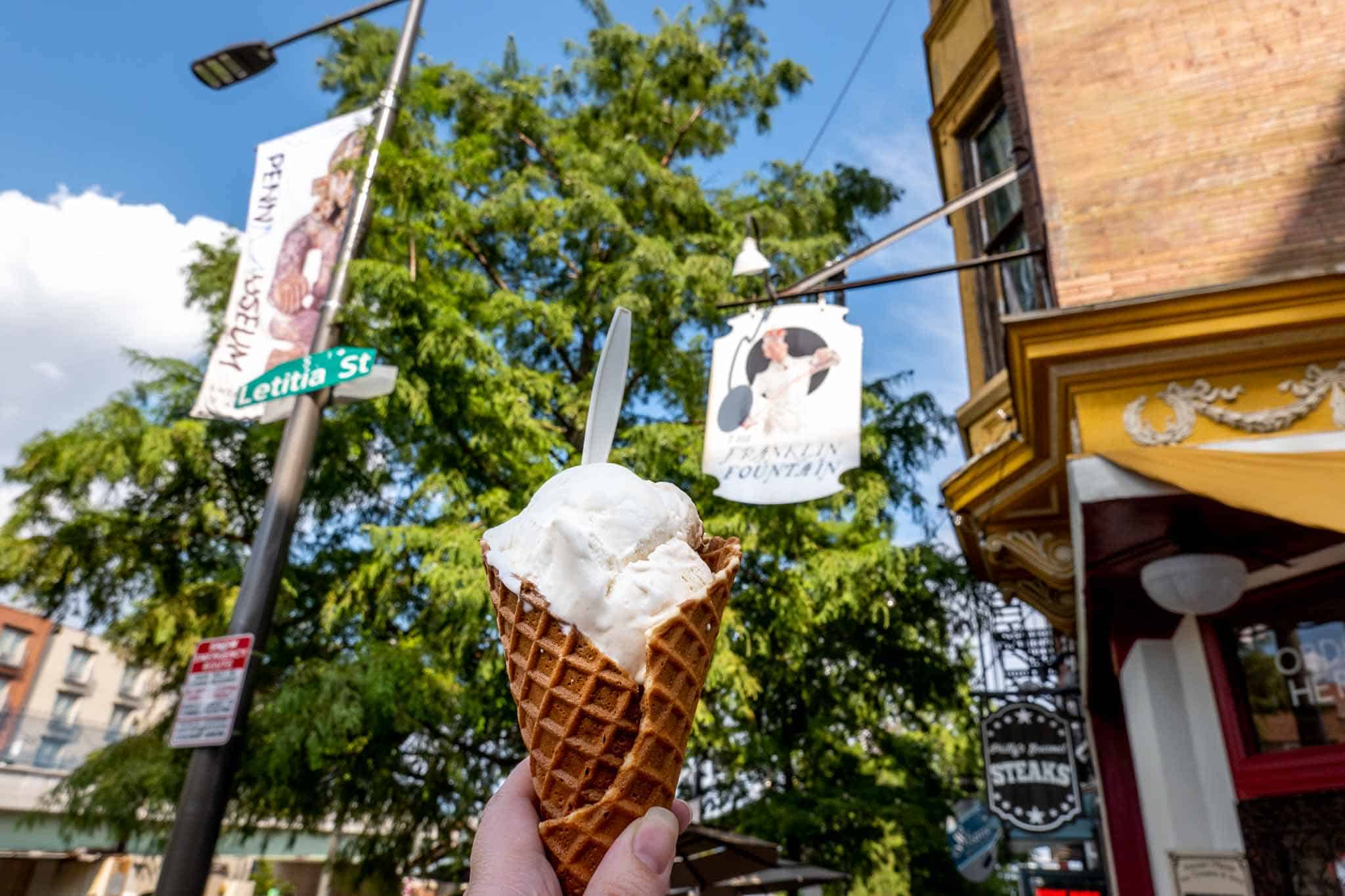 Hand holding an ice cream cone in front of a sign for The Franklin Fountain.