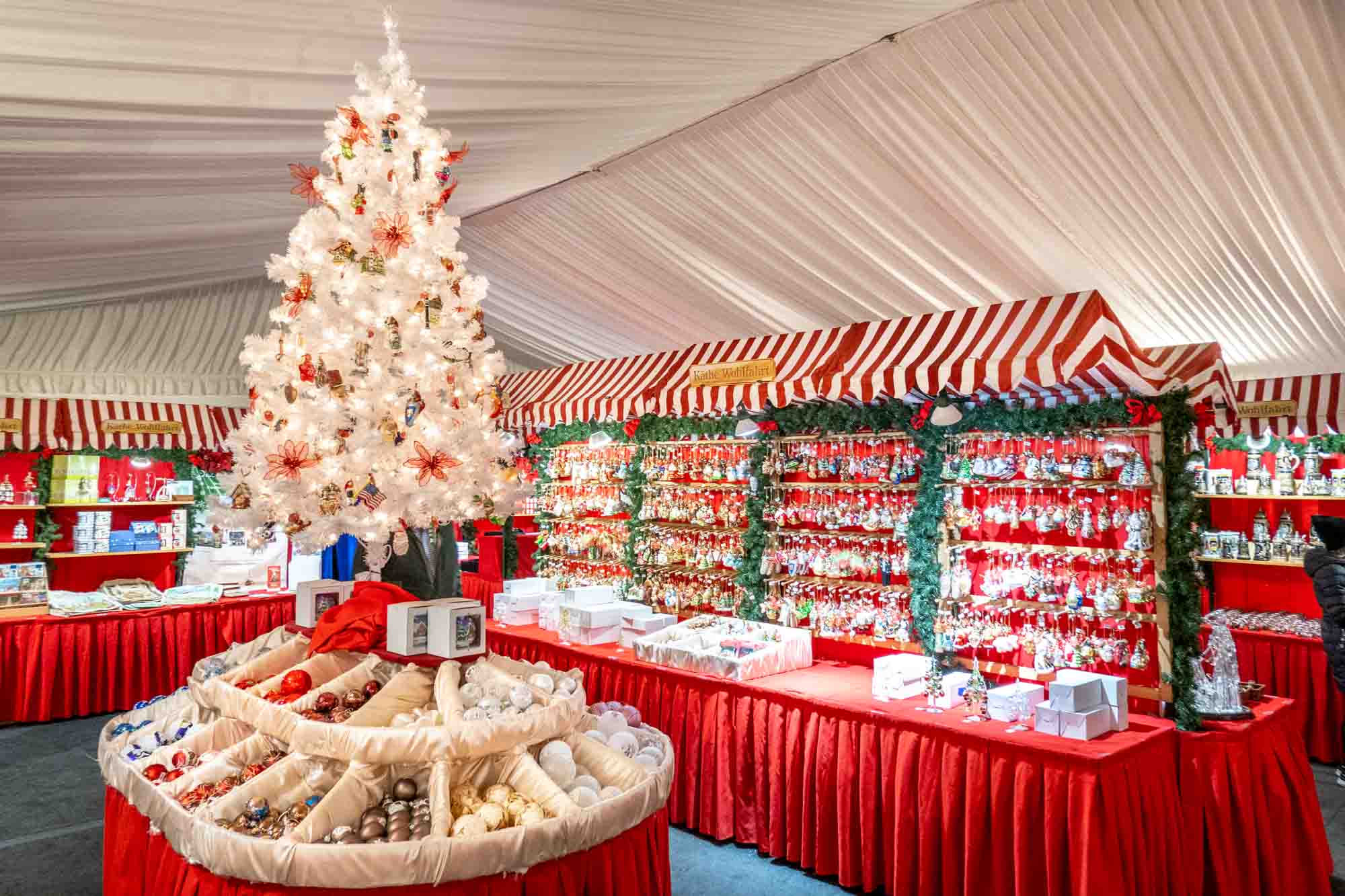 Rows of hanging ornaments for sale and a white Christmas tree under a tent.