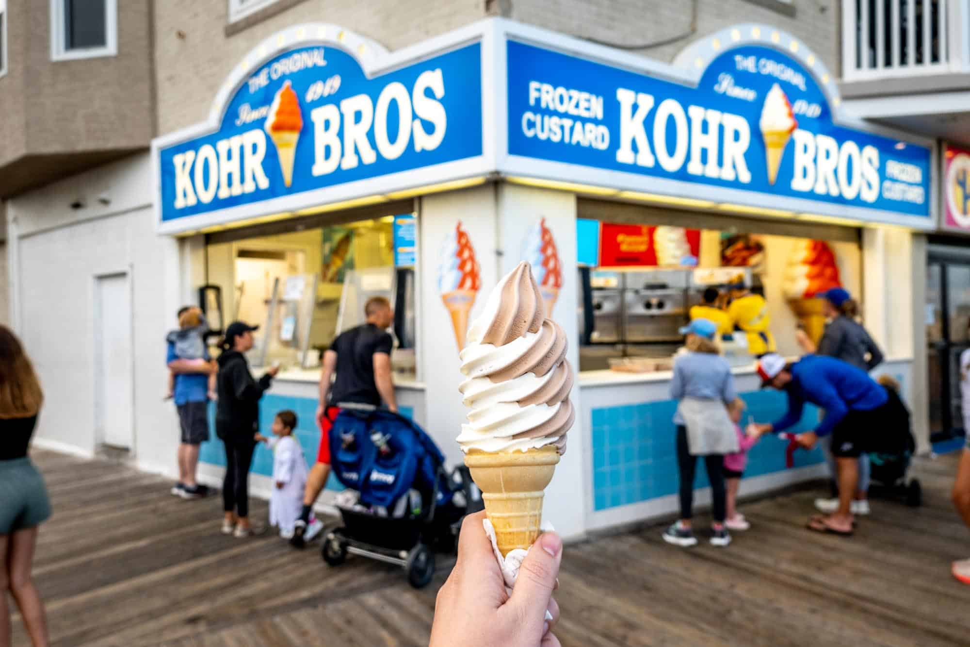 Hand holidng a cone of frozen custard in front of Kohr Bros store.