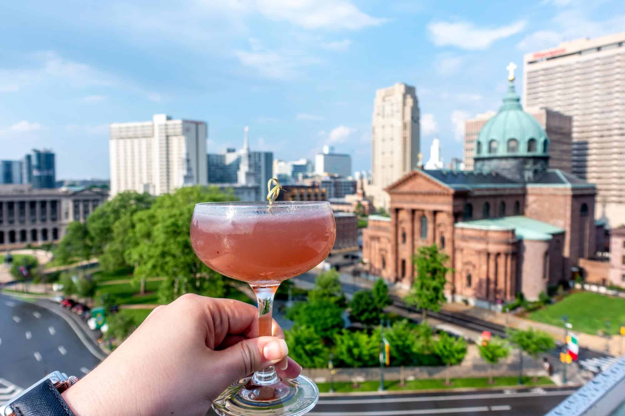 Hand holding a cocktail glass in front of a view of a park and ornate building below.