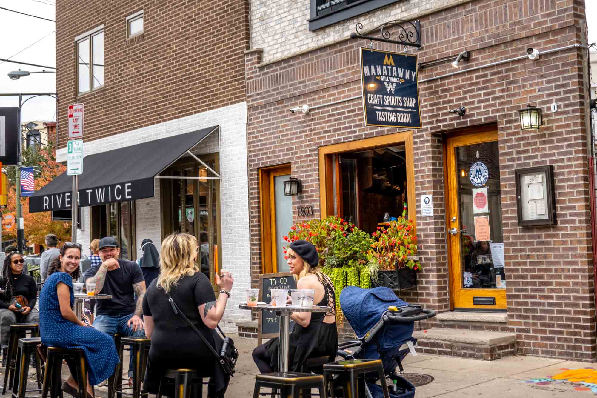 People seated at cocktail tables outside a brick building with a sign for "Manatawny Still Works: Craft Spirits Shop, Tasting Room"