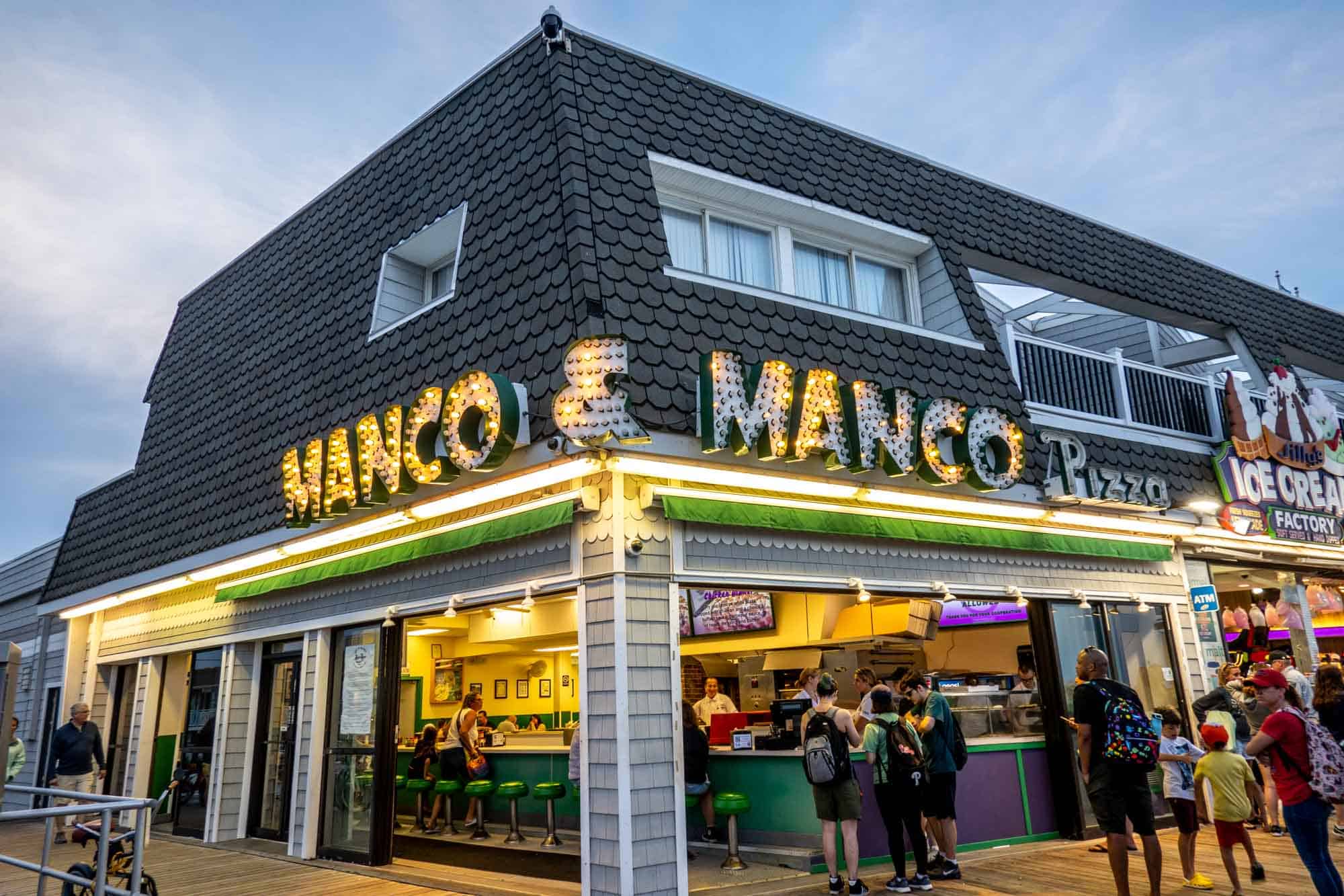 People in a restaurant with open garage-style doors below a sign for "Manco & Manco Pizza."