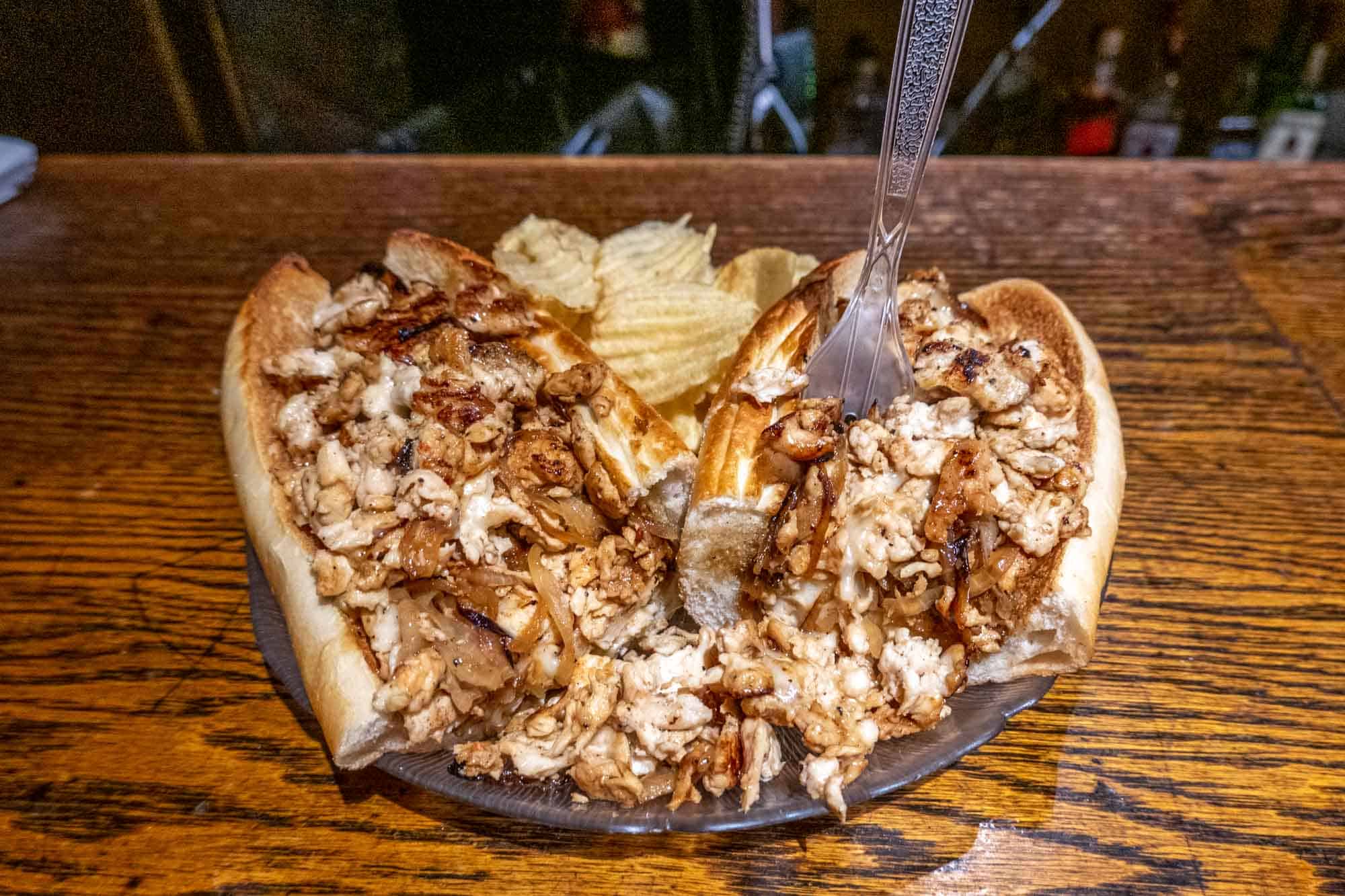 Chicken cheesesteak with a plastic fork and potato chips on a plate.