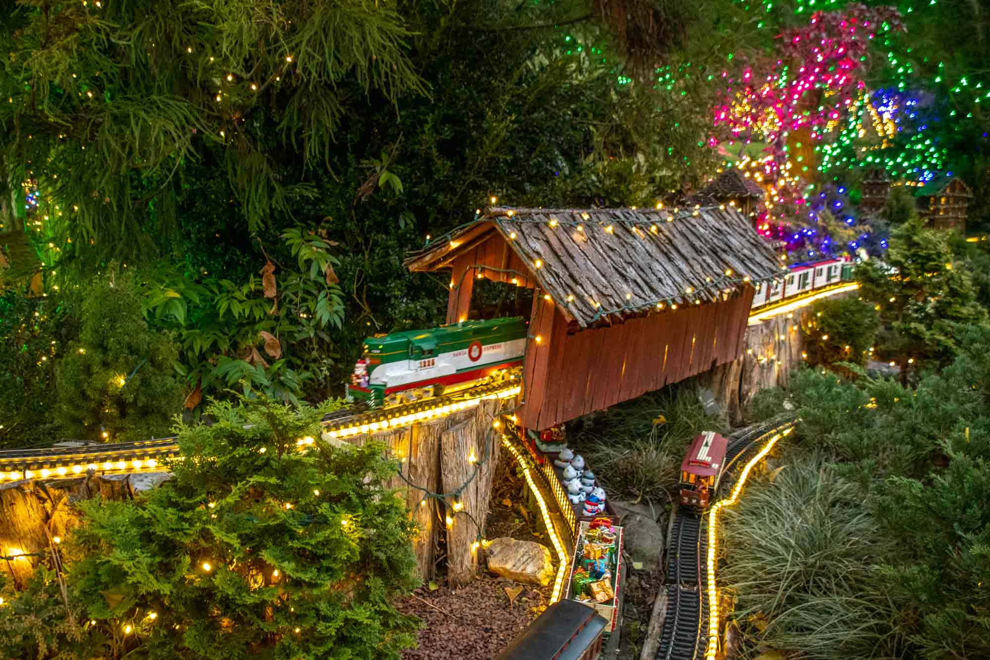 Model train crossing a covered bridge in an environment lit with Christmas lights.