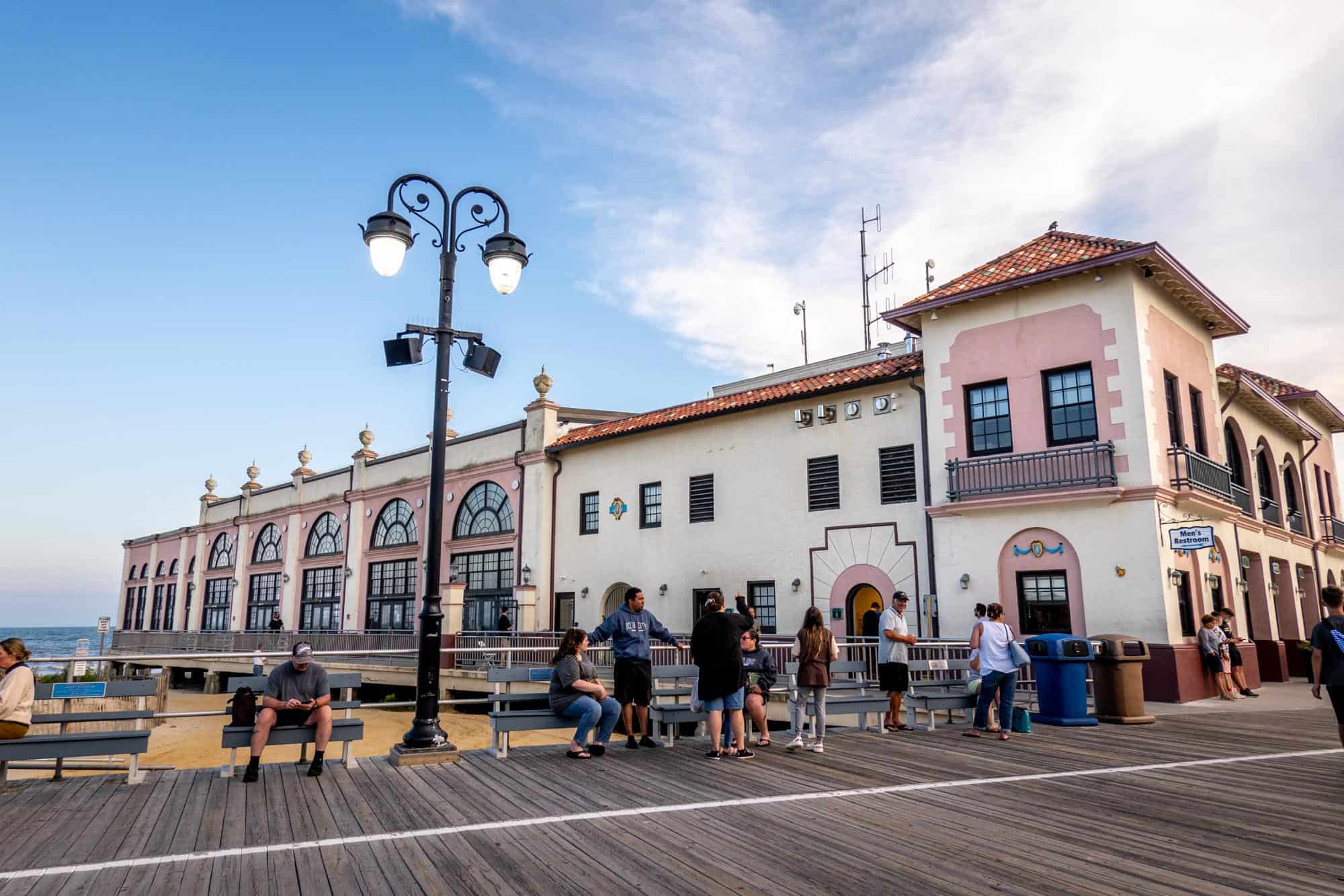 People beside a white building with pink accents and arched windows just off the boardwalk.