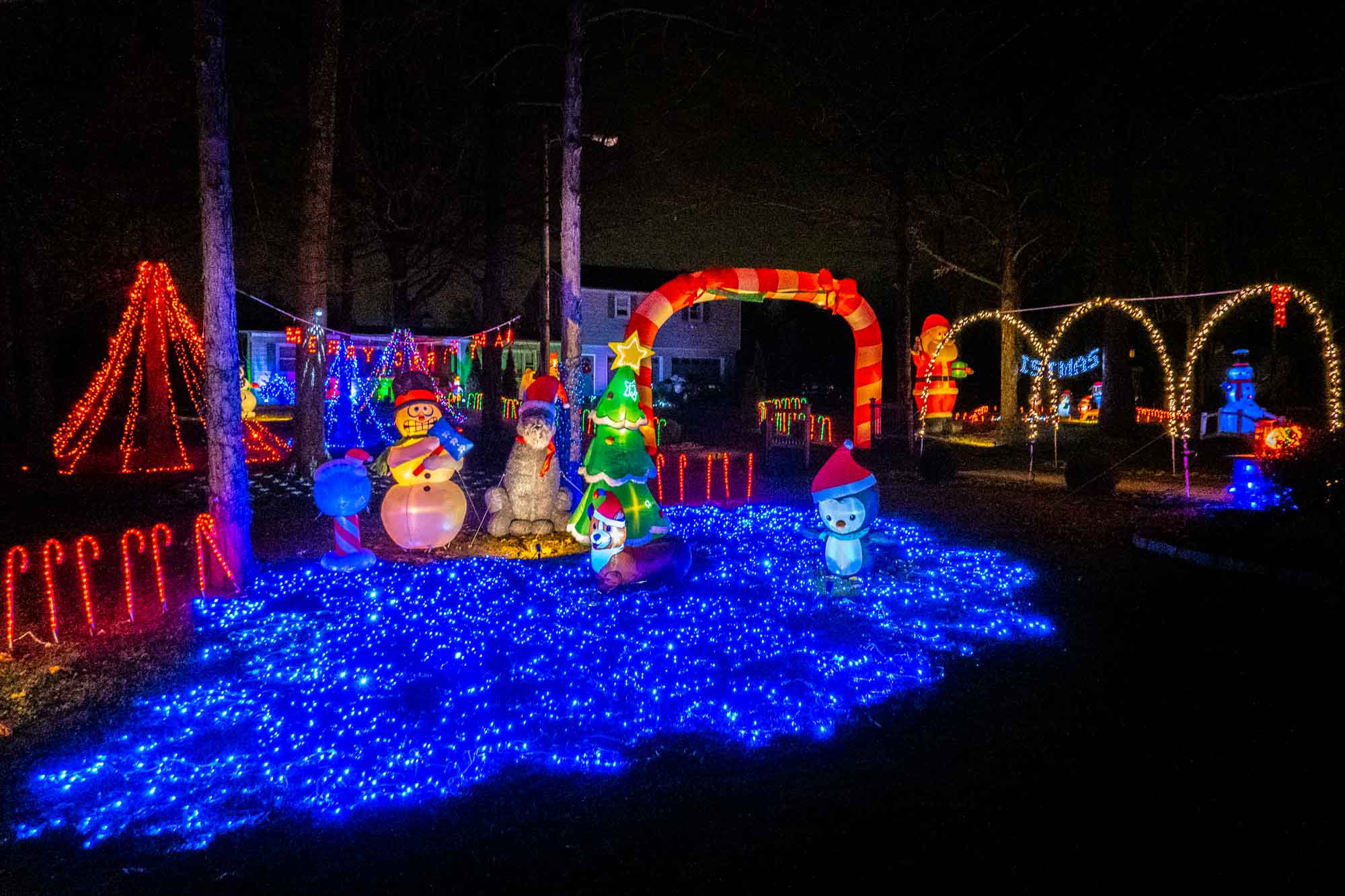 Christmas light display including arches, trees, and an inflatable snowman at night.