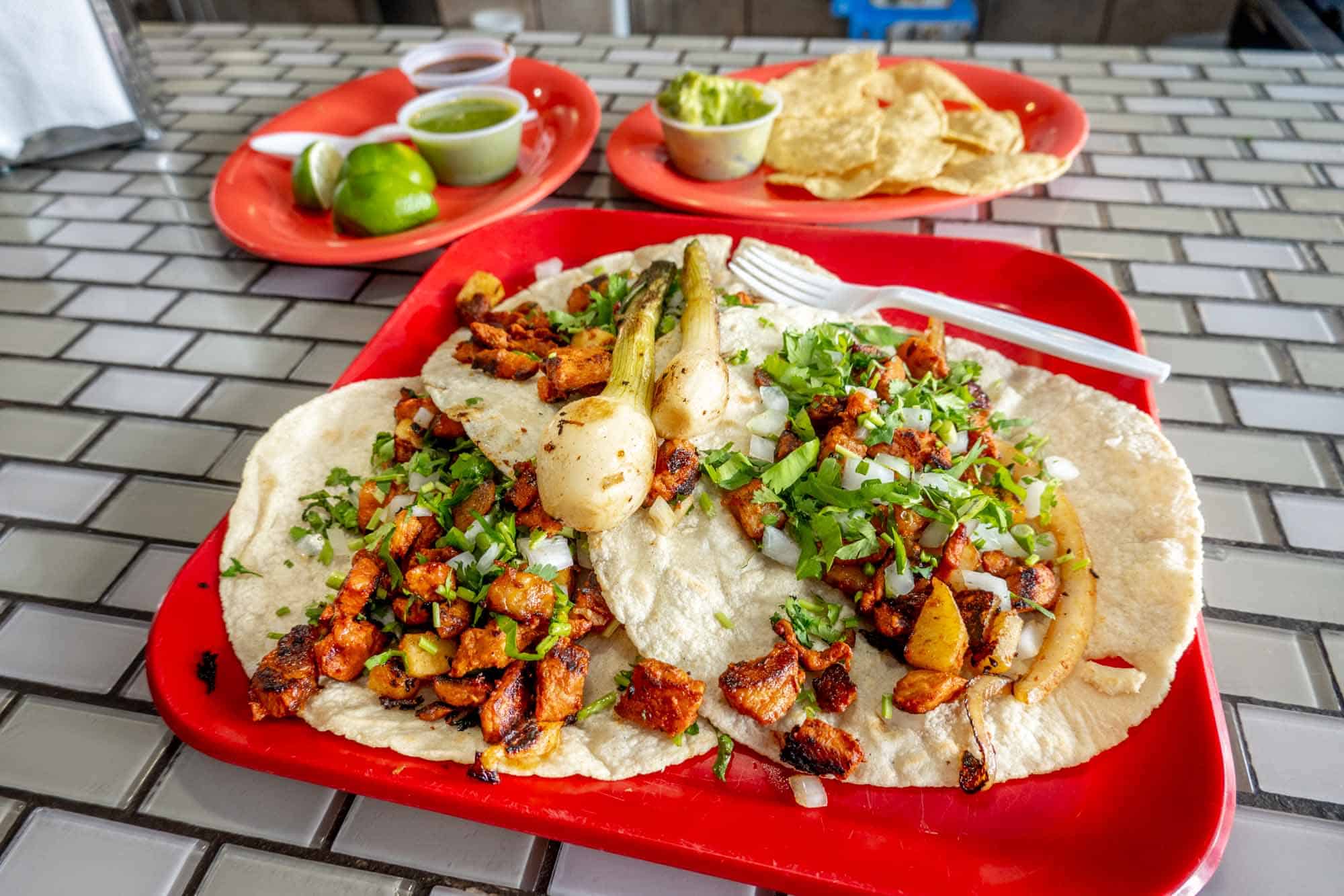 Tacos al pastor on a red try beside other trays containing tortilla chips, guacamole, limes, and salsa.