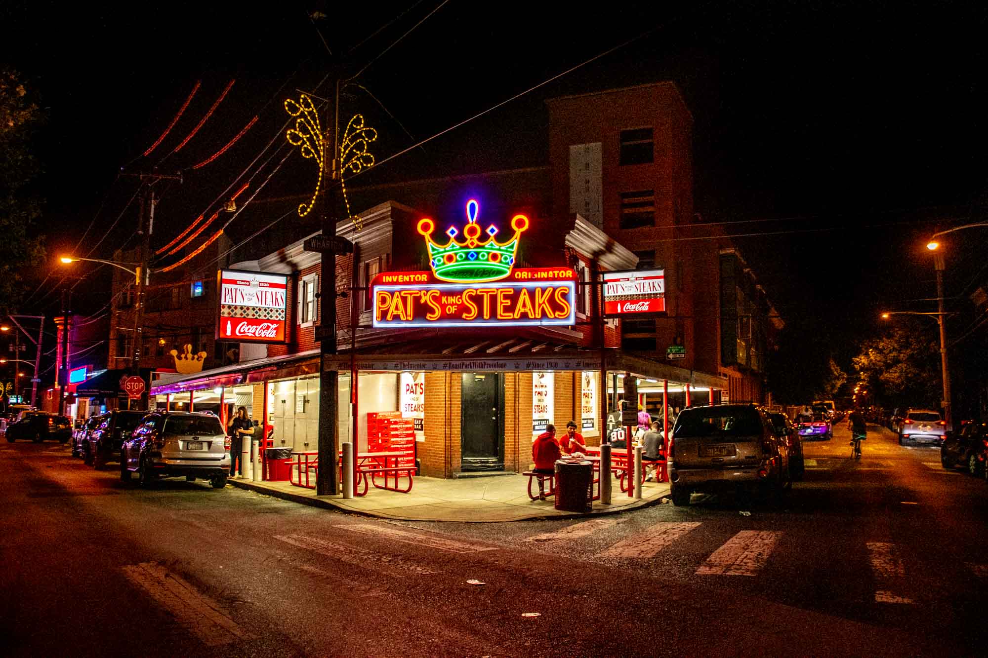 Exterior of a brick restaurant at night with a neon sign saying "Pat's King of Steaks."