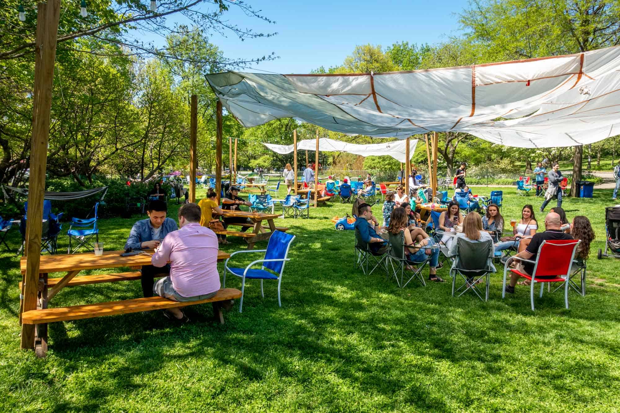 People sitting at picnic tables and in chairs at an outdoor beer garden in a park.