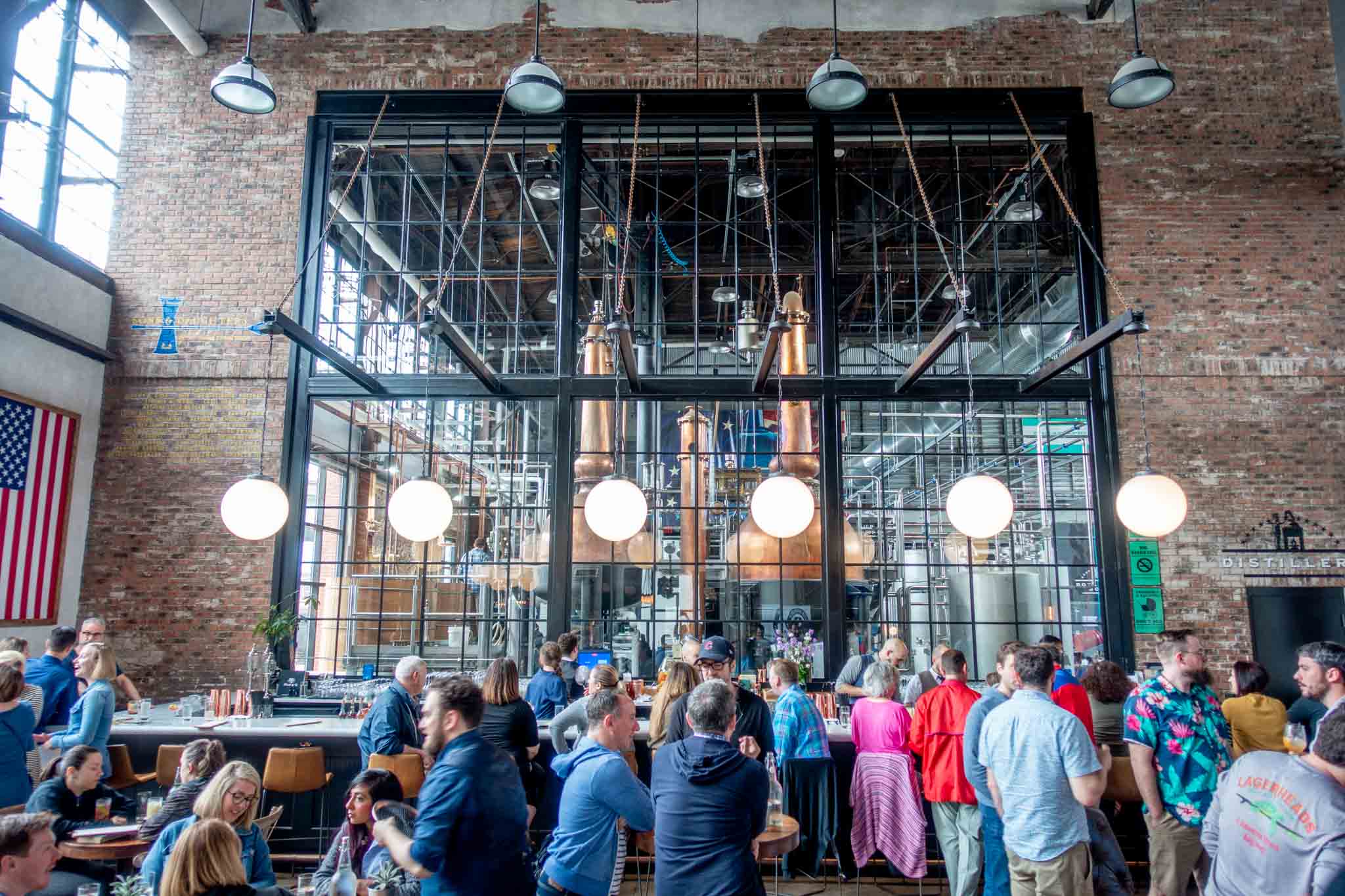 People standing at a bar in front of a huge window with large copper stills on the other side