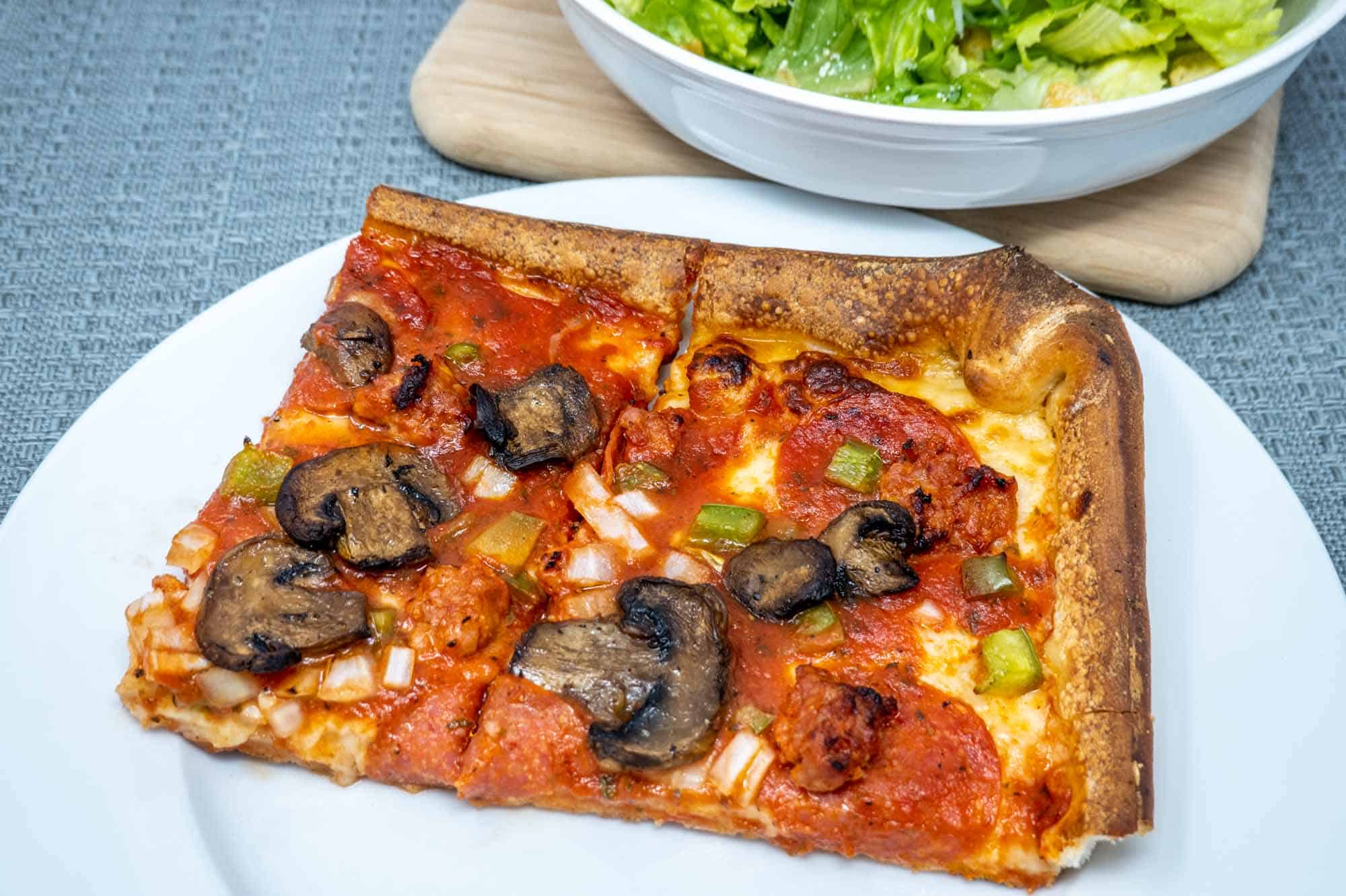 Two square pizza slices with pepperoni, mushroom, onion and green pepper on a plate beside a bowl of salad.