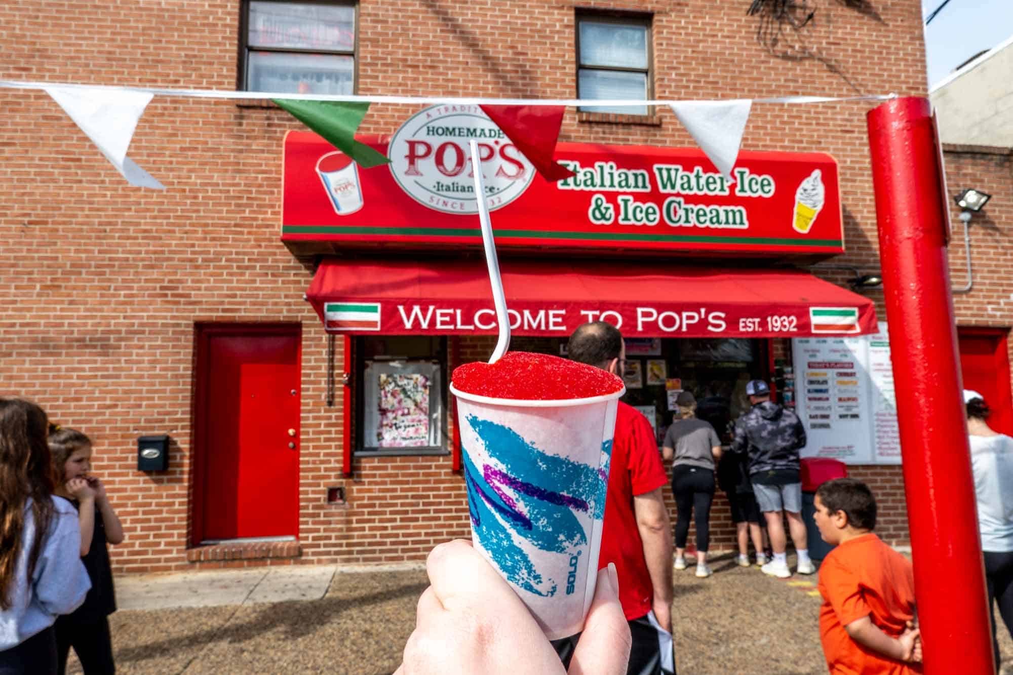 Hand holding a cup of red water ice in front of people waiting in line at a business with a red awning: "Pop's Italian Water Ice & Ice Cream."