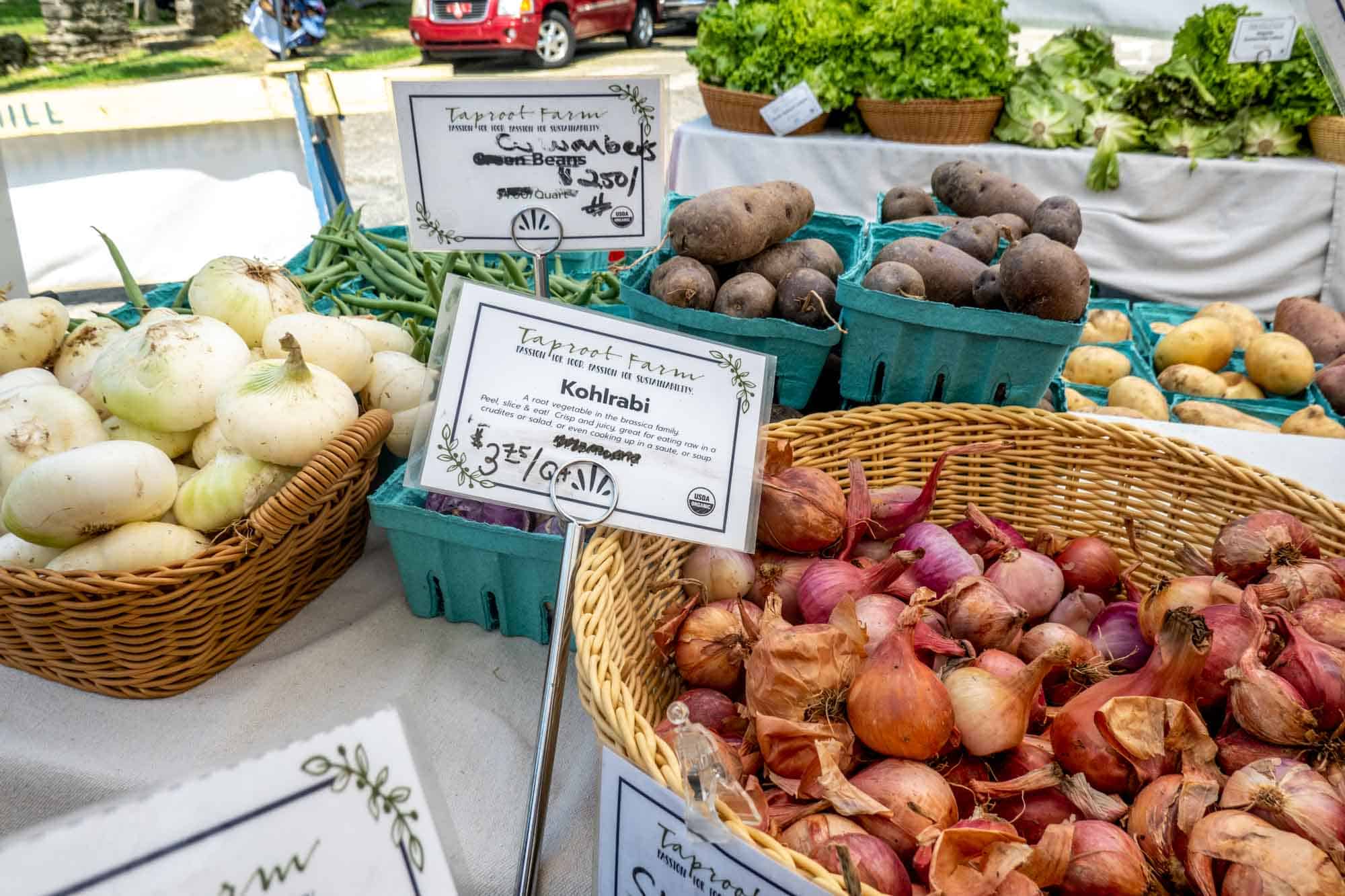 Wicker baskets of shallots and kohlrabi, along with containers of purple potatoes and yellow gold potatoes