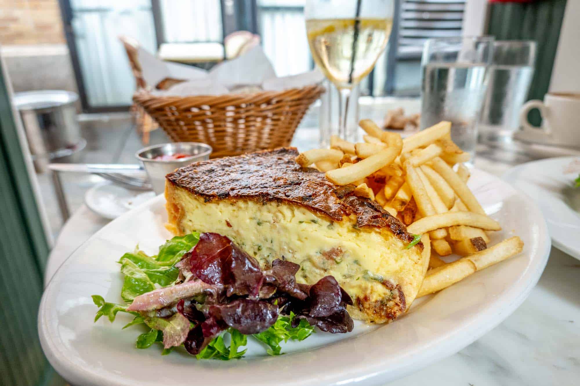 Slice of quiche on a plate with French fries and a salad and a cocktail and basket in the background.